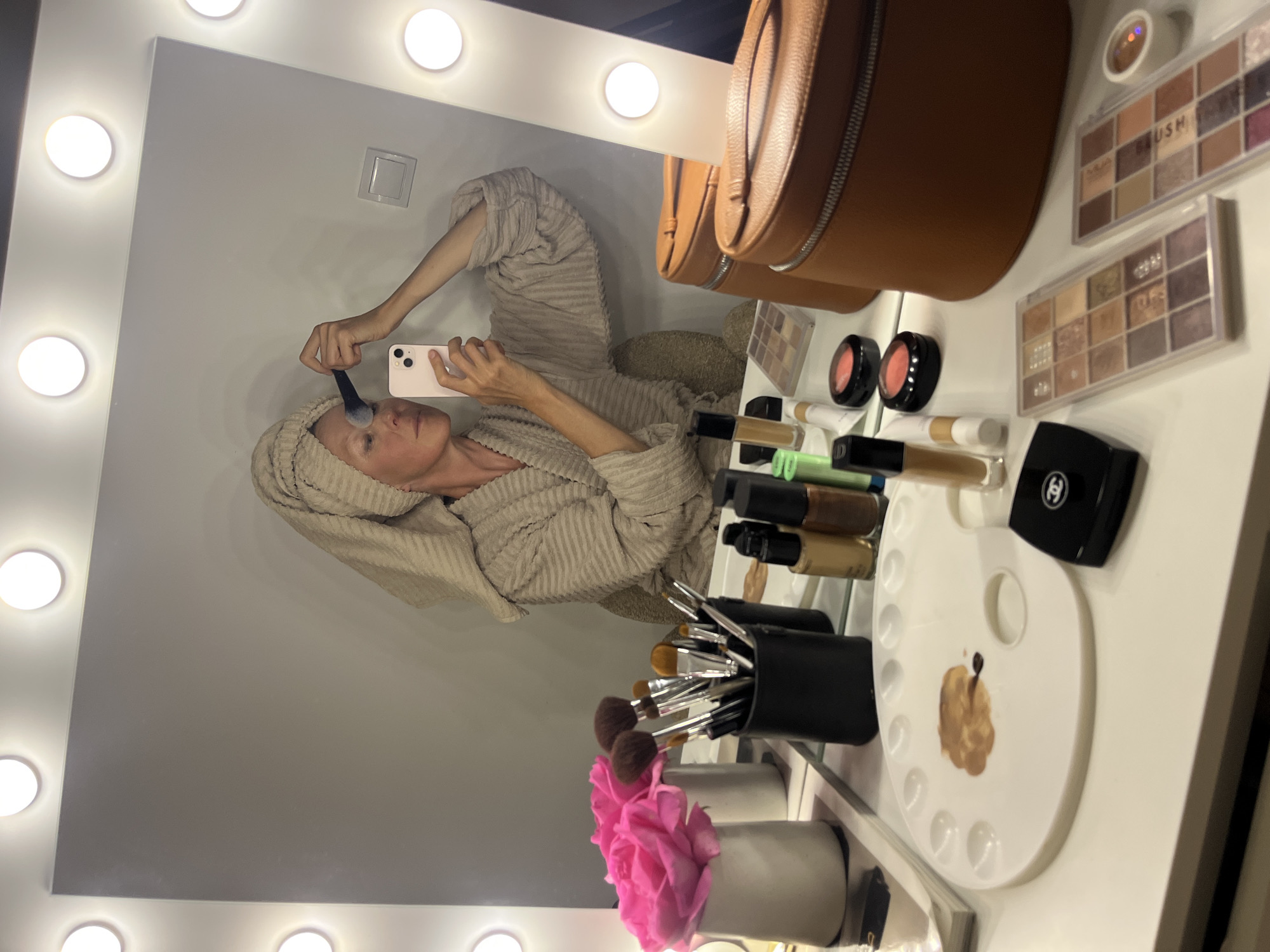 The image depicts a mirror selfie of a woman with a towel wrapped around her head. She's wearing a robe and applying makeup with a brush while holding a smartphone. The selfie is taken in front of a vanity mirror surrounded by lights. On the vanity surface are various makeup items, including eyeshadow palettes, blush compacts, foundation bottles, a makeup brush holder filled with brushes, a white palette with makeup on it, and a Chanel compact. A pink floral arrangement sits in front of the makeup brushes. The scene does not provide information to locate this scene in the city of Budva, Montenegro.