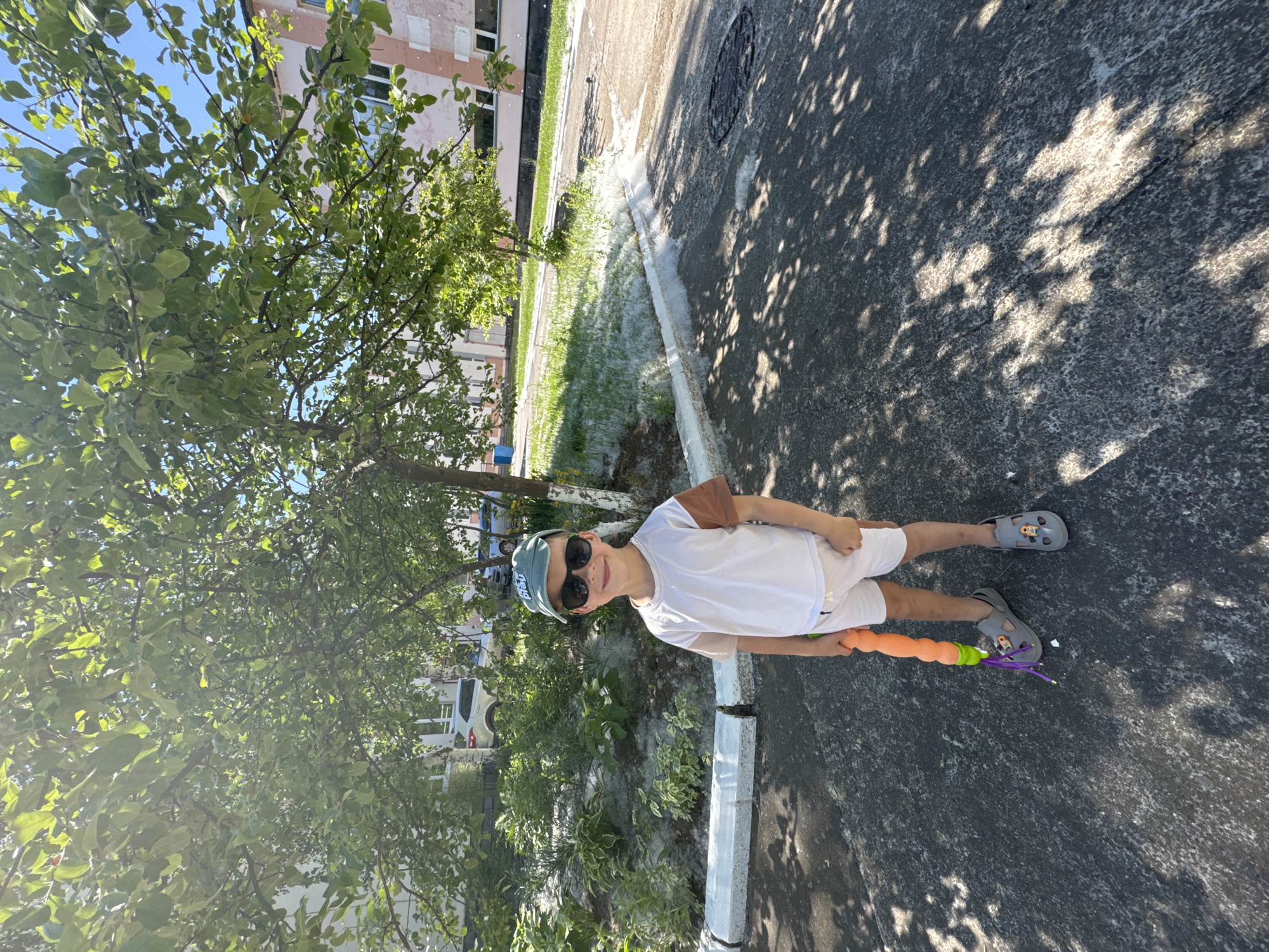 A young boy stands on an asphalt surface in front of a garden bed covered in white fluff. He is wearing white shorts, a white t-shirt with brown sleeves, a green baseball cap, and sunglasses. He holds an orange toy with purple streamers in his right hand. The boy is positioned in front of a small tree with green leaves. In the background, there are buildings with pink and white facades, parked cars, and more green trees. The ground and garden bed are covered in a significant amount of white fluff, likely poplar or cottonwood down, indicating a springtime or early summer scene. The lighting suggests it is a sunny day, with dappled sunlight casting shadows on the asphalt. The location is identified as Chaikovsky, Russia, likely a residential area given the presence of apartment buildings and a garden bed.