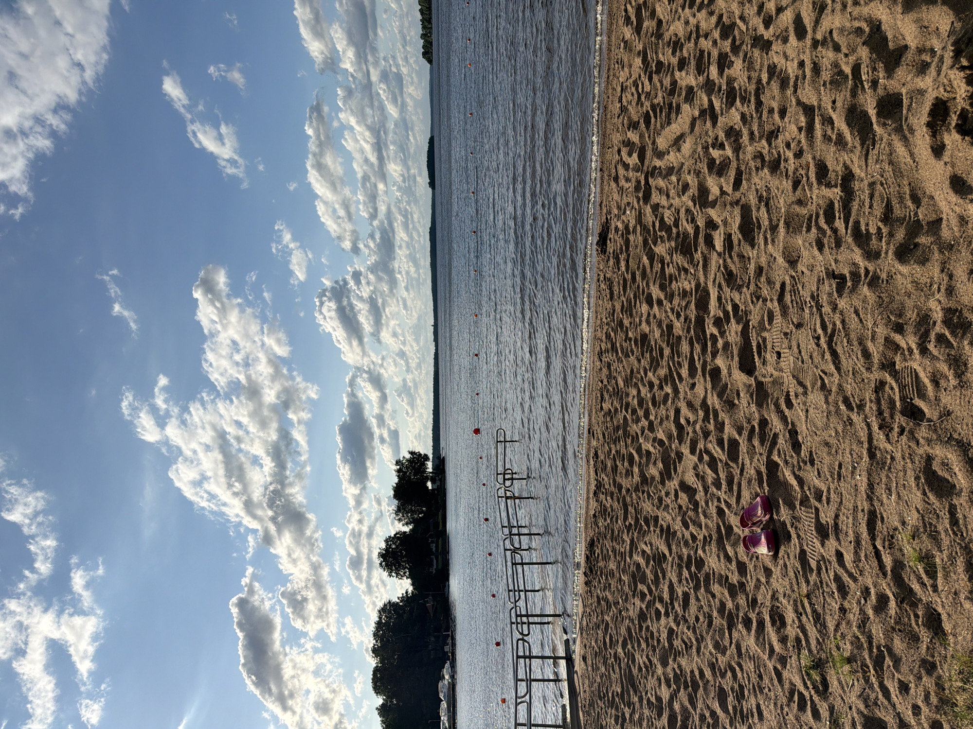 A tranquil scene unfolds at the sandy beach of a lake in Växjö, Sweden. The foreground is dominated by the texture of fine, light brown sand, showing the imprint of footprints and subtle undulations. A pair of pink flip-flops rests side-by-side on the sand, about a third of the way up from the bottom edge of the image. Beyond the beach, a calm, dark blue lake stretches out to the horizon. A metal pier with handrails extends into the water from the left side of the frame. Along the waterline, a series of orange buoys are spaced out in a line. In the middle ground, the far shore of the lake is lined with a dense growth of green trees, with a few buildings and a cluster of white boats docked on the left. The sky above is a brilliant blue, dotted with large, fluffy white clouds, some catching the sunlight and appearing luminous. The overall impression is one of peaceful, sunny weather at a lakeside recreational area.