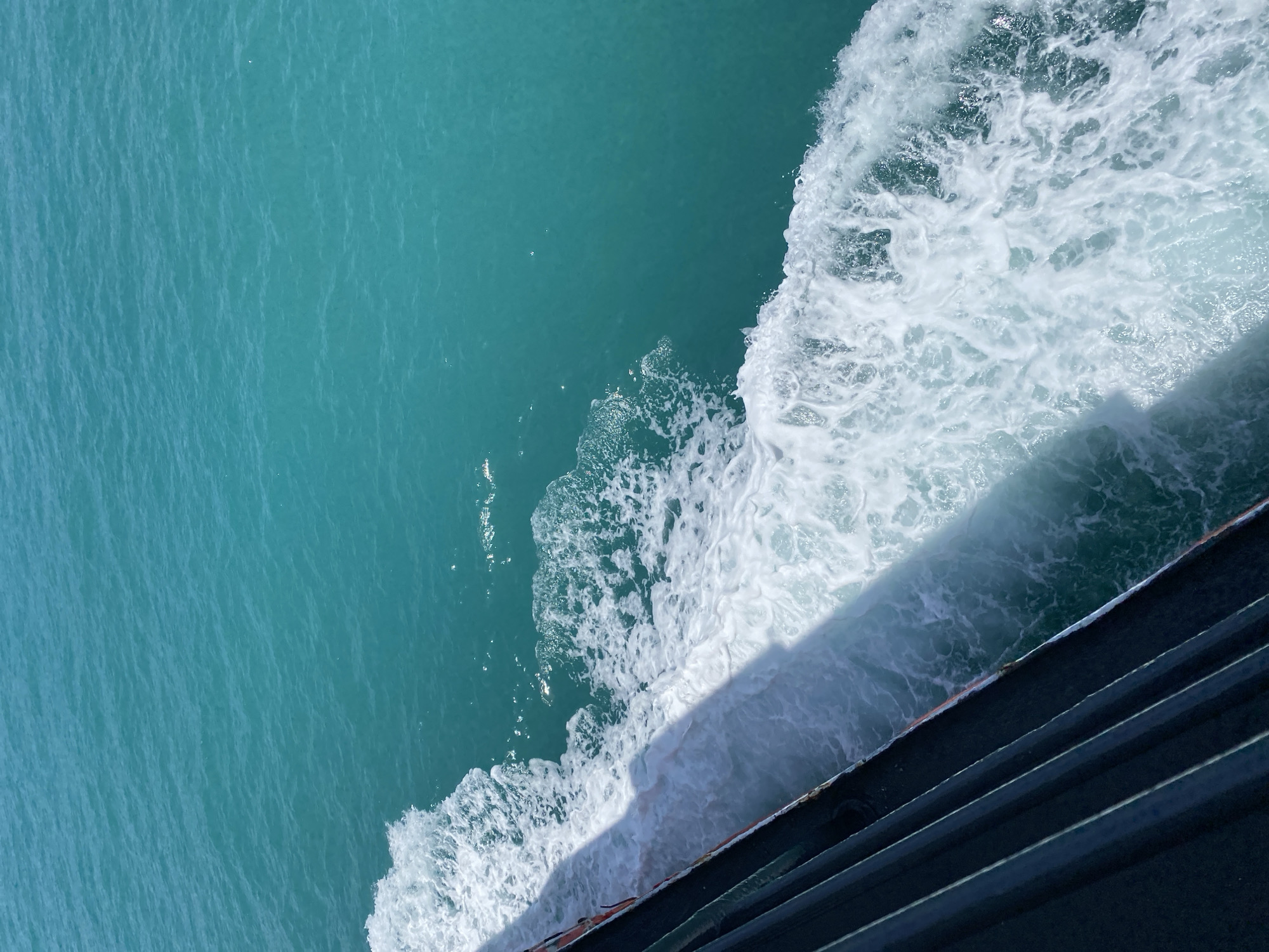 This is a high-angle, close-up shot of turquoise water with white foam, viewed from the side of a vessel. The dark hull of the vessel is visible in the lower left quadrant of the image, with its shadow cast across the turbulent water. The water appears to be in motion, creating a wake. The scene suggests a journey at sea, possibly a ferry or boat ride. The bright sunlight on the water indicates a clear day. The specific location of Ko Samui District, Thailand, implies a tropical or subtropical setting, consistent with the vibrant blue water. There are no people visible in the frame.
