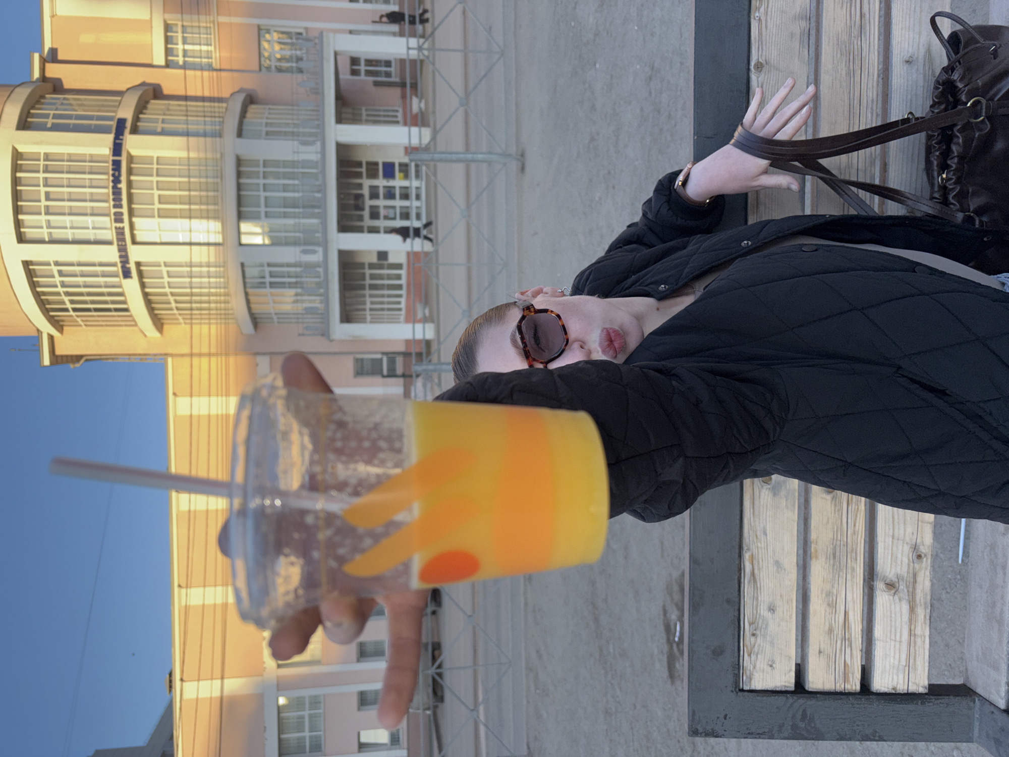 A person is seated on a bench in Perm, Russia, holding a cup of orange liquid. The person is wearing sunglasses and a black quilted jacket. A black bag is next to the person on the bench, which is made of wooden slats. The background shows a light-colored building with multiple windows. The cup contains a straw and has orange decorations. The sky is blue.