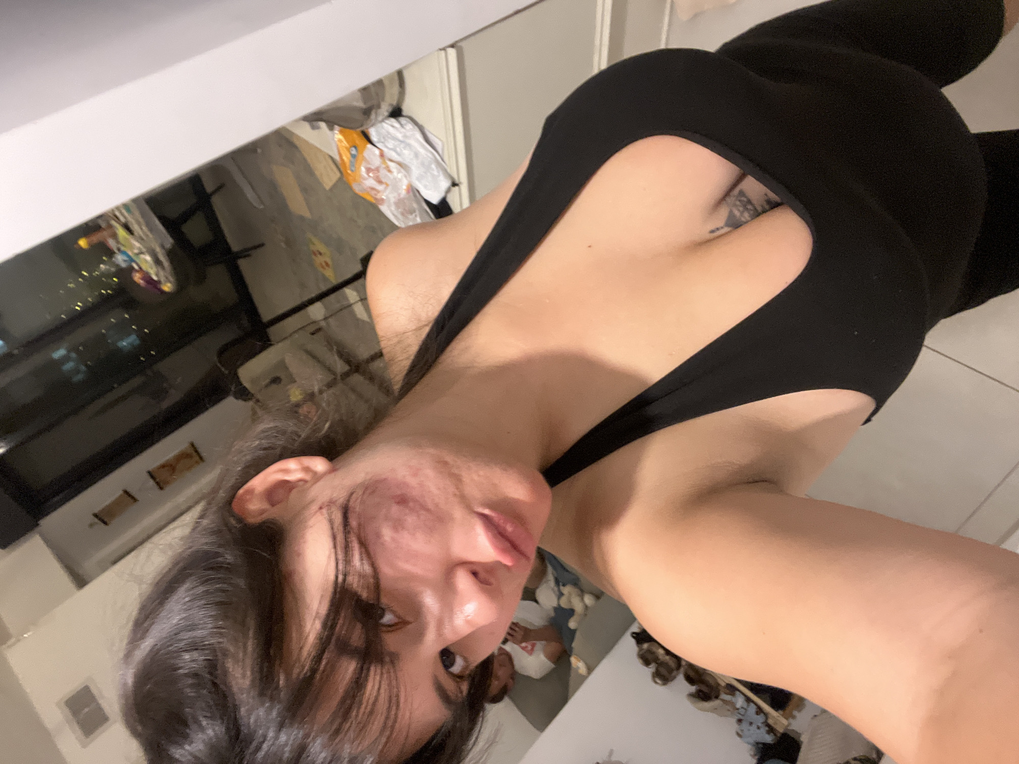 The image shows a woman in a black tank top, with dark hair pulled back. She is looking at the camera, with her mouth slightly open. She has acne on her face. In the background, there is a person sitting on a couch, looking at their phone. There is also a table with items on it, and a window with a view of a city at night. The setting appears to be an apartment. The image does not contain any specific landmarks or indicators to identify its location in Kiryat Bialik, Israel.
