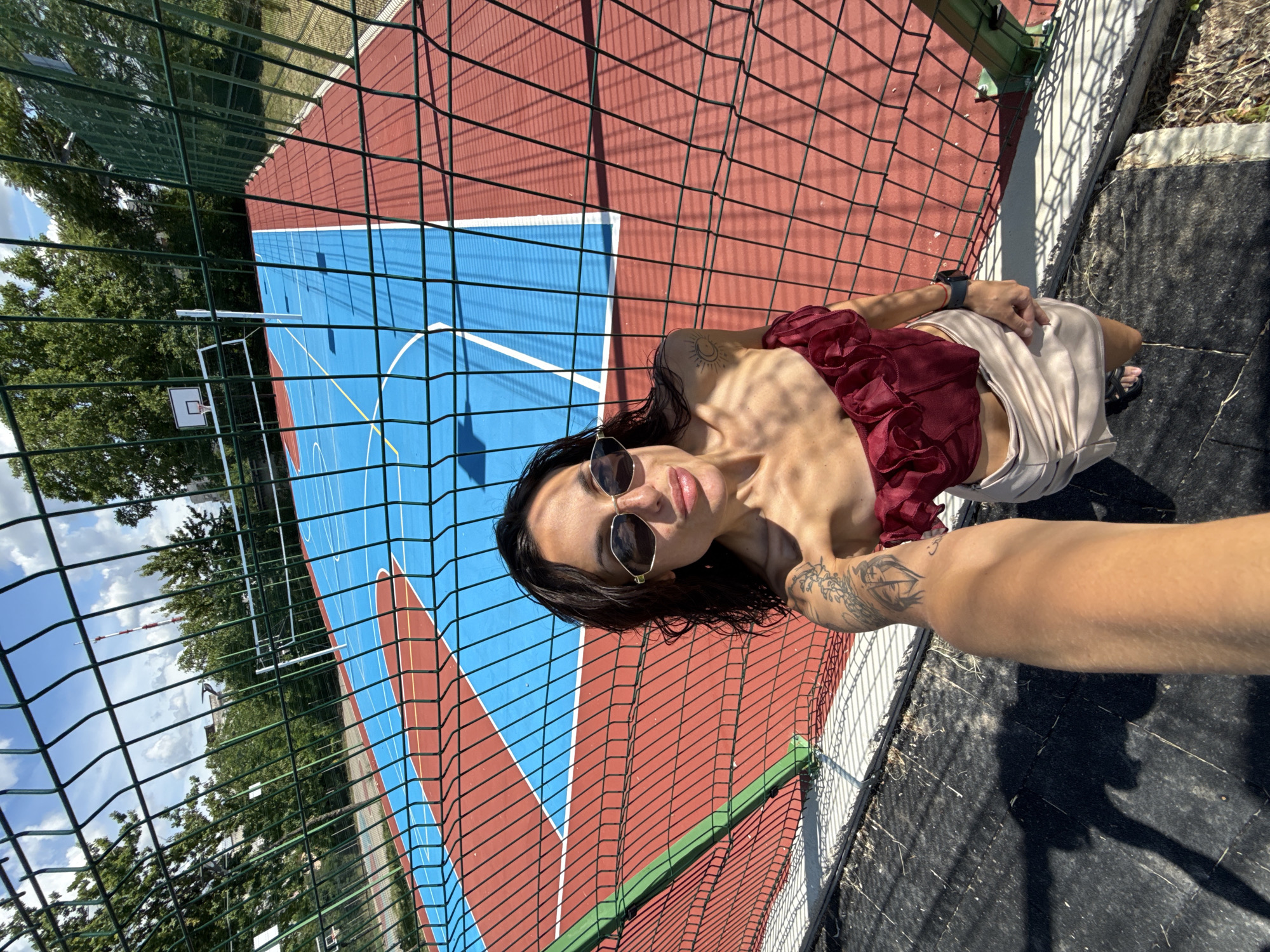A woman takes a selfie on a sunny day at an outdoor sports complex. She is wearing a maroon ruffled top and a light-colored skirt, with sunglasses and noticeable tattoos on her arm and shoulder. Behind her, a blue and red basketball court is visible, complete with markings and a net. A tall chain-link fence separates her from the court. The sky is blue with scattered clouds. The sports complex appears to be located in an urban area, with trees and buildings visible in the background, suggesting it could be a public park or recreational facility in Pleven, Bulgaria.
