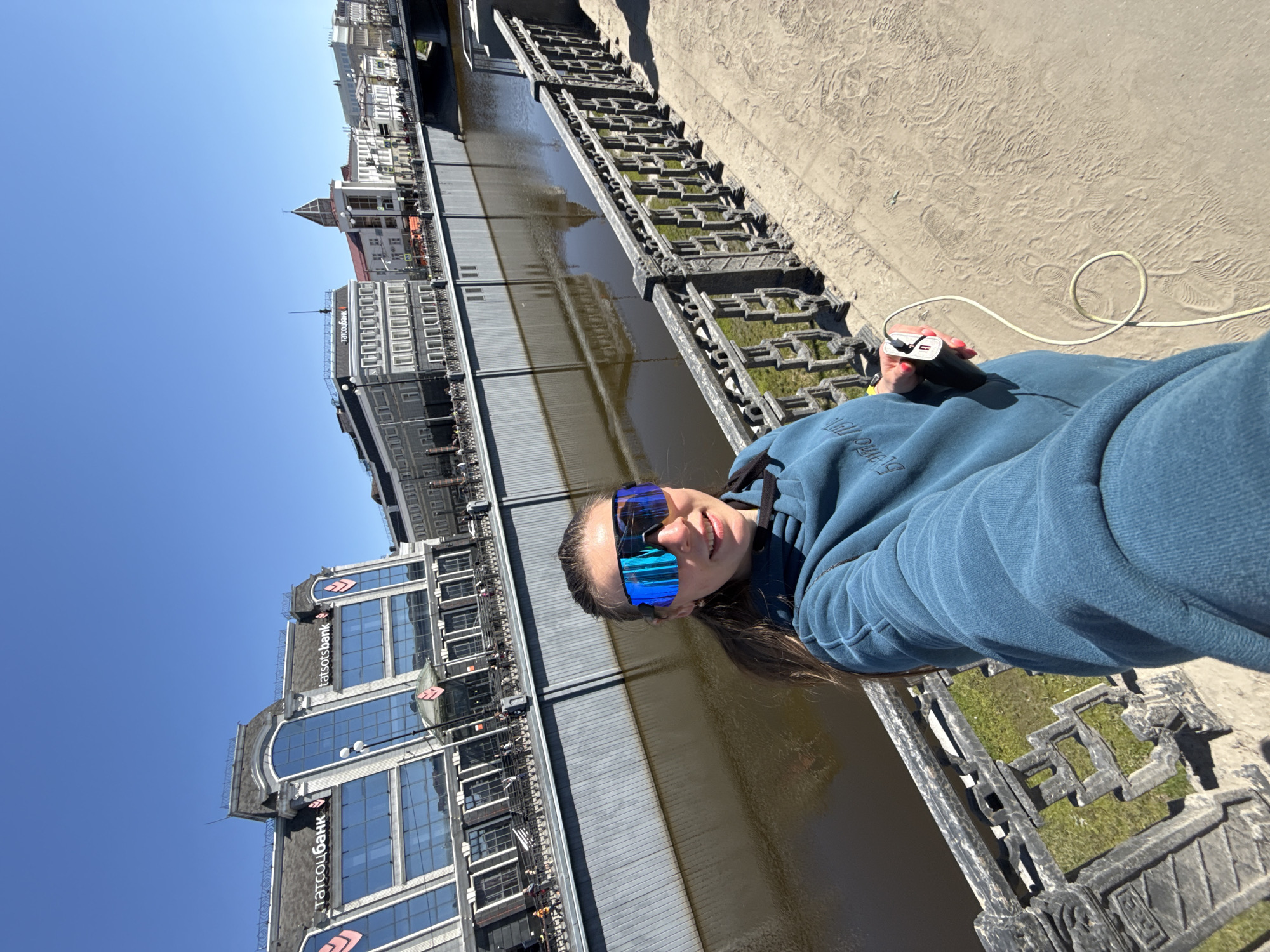 A person is holding a phone charger while standing next to a river in Kazan, Russia. The river's edge is lined with ornate, dark gray fencing. Buildings are visible on the opposite side of the river, including a building marked with "татсоцбанк" and "tatotsbank." The sky is clear blue. The person is wearing blue reflective sunglasses and a blue hoodie. The person is holding a black phone charger with a white cable. The ground next to the river is light brown.