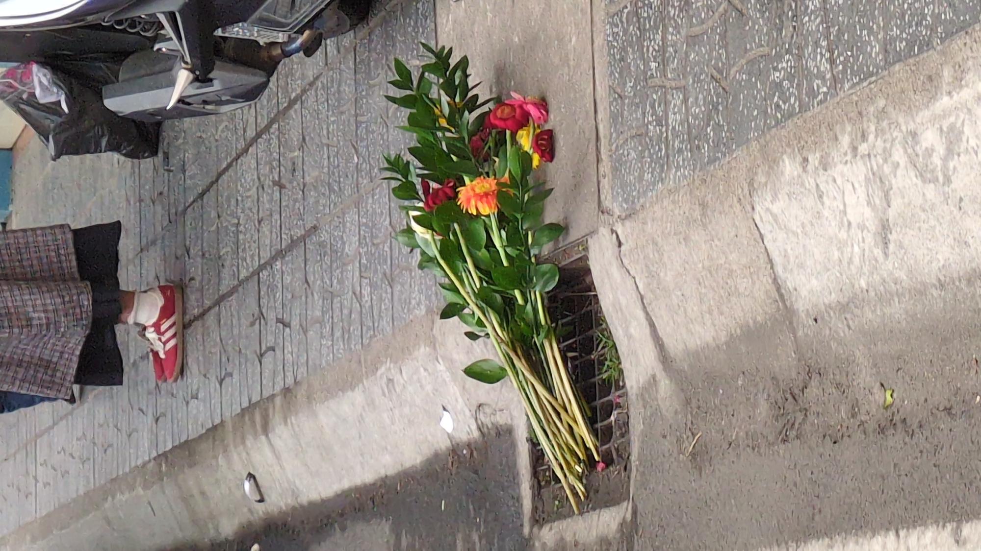 The image shows a bundle of flowers discarded in a storm drain next to a sidewalk in what may be Da Lat, Vietnam. The flowers are a mix of yellow, orange, and red colors, with green stems and leaves. To the side of the flowers, there is a foot wearing a red Adidas shoe, belonging to an individual partially visible in the image, wearing black pants and a brown and gray plaid jacket. The ground appears to be made of concrete. A motorbike is parked on the sidewalk to the left of the foot. The storm drain is constructed of iron bars.