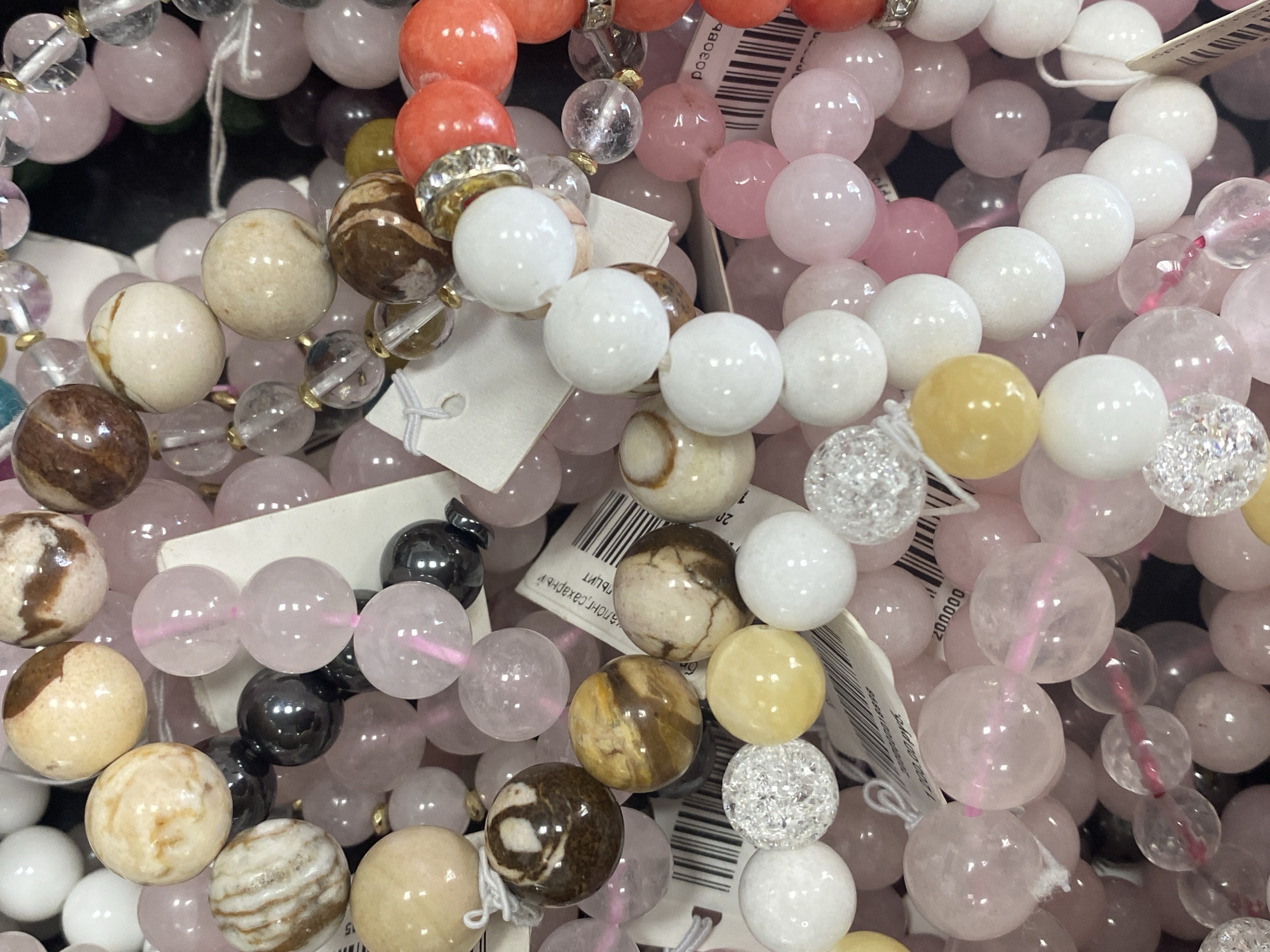 The image shows a close-up of several beaded bracelets, possibly for sale, likely taken in the city of Yekaterinburg, Russia based on visible Cyrillic text on a tag. The bracelets consist of various types of beads, including white, pink, orange, brown, clear, and yellow spherical beads, as well as some beads with a crackled appearance. Some of the bracelets have tags attached. The beads are strung together with what appears to be elastic cords. The bracelets are clustered together, suggesting they are displayed in a shop or market setting.