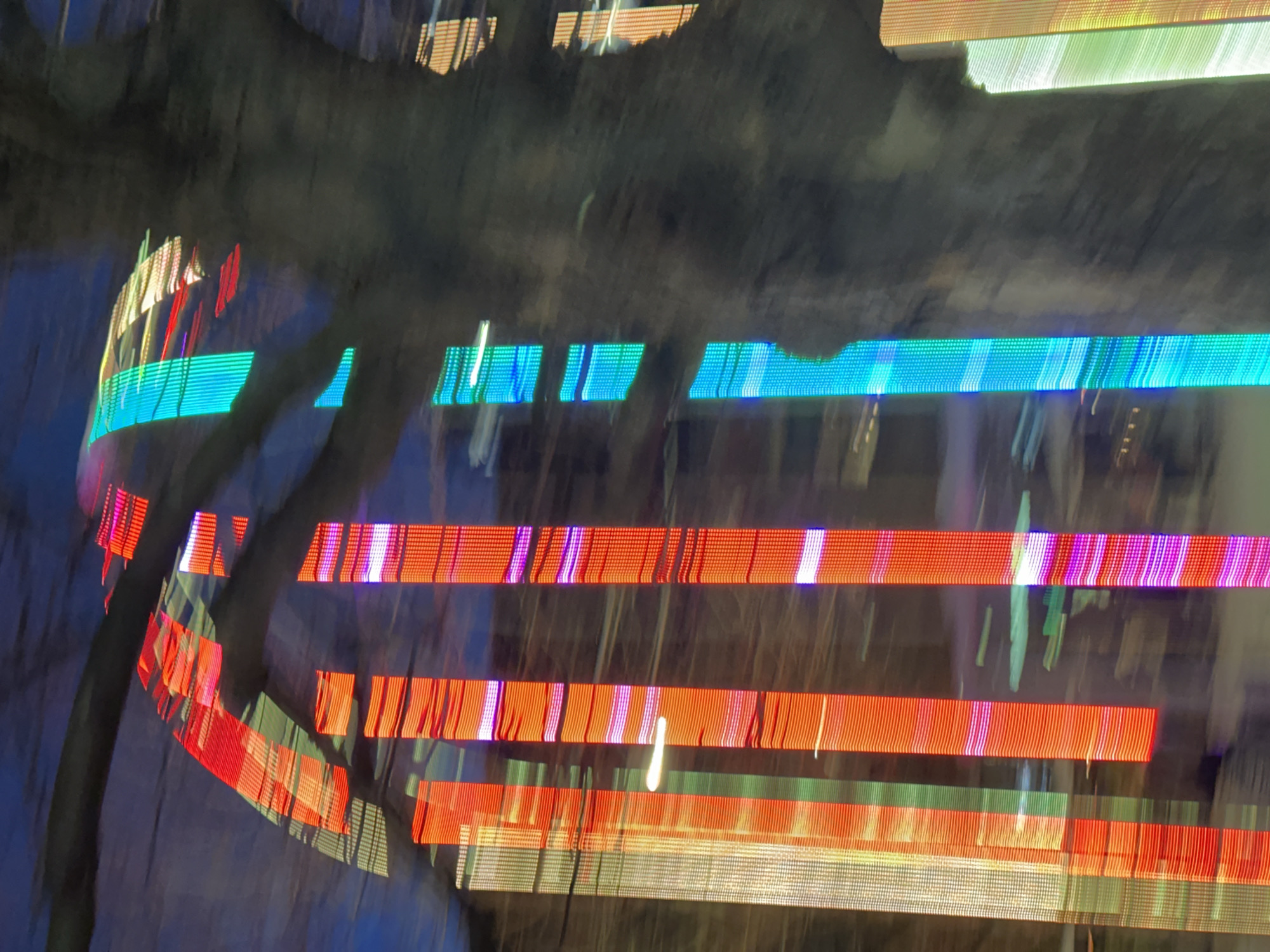 This image presents a highly blurred, vertical composition dominated by vibrant, elongated light streaks and a prominent dark, organic mass. Numerous vertical bands of light in colors including red, white, green, blue, orange, and magenta traverse the frame, suggesting reflections or illuminated digital displays. These light columns exhibit a pixellated or segmented quality. In the upper left, a distinct arch structure, primarily illuminated in red, is discernible, characteristic of Tower Bridge. A large, dark, textured, vertical mass, consistent with a tree trunk or thick pillar, occupies the right side of the image and extends diagonally into the upper frame, partially obscuring the lights. The severe motion blur and light streaking indicate either significant camera movement, reflections from a moving surface, or a long-exposure photograph. The scene is located in London, United Kingdom, specifically depicting or reflecting parts of Tower Bridge and adjacent illuminated urban architecture along the River Thames. No individuals or specific actions are identifiable due to the image's abstract nature.