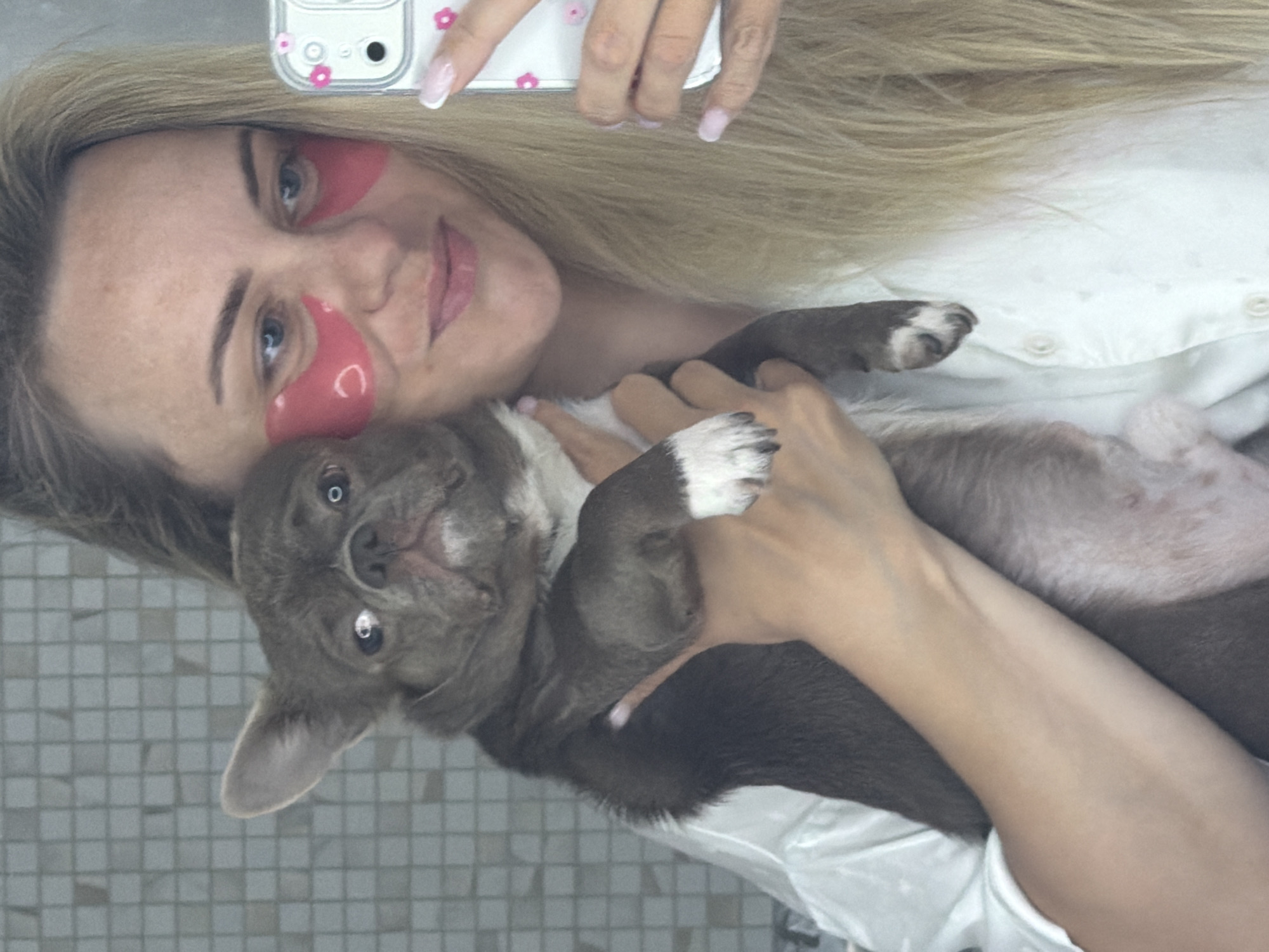 A blonde woman takes a selfie in an indoor setting in Votkinsk, Russia, holding a small grey-brown dog, likely a chihuahua or similar breed. She is seen from the chest up, with a slight smile on her face. She has pink under-eye patches applied, suggesting a self-care routine. Her long blonde hair frames her face.
The woman holds a white smartphone with a transparent case adorned with pink flower stickers, visible in the upper right corner of the frame. Her manicured nails, some with white French tips and others with pink details, are also visible as she grips the phone. She is wearing a white collared shirt with buttons.
The dog is cradled in her arms, looking upwards towards the camera with a curious or relaxed expression. Its white-marked paws are visible. The background features light grey square tiles, indicating an indoor environment such as a bathroom or a room with tiled walls. The lighting is bright, consistent with an indoor daytime or well-lit evening setting. There is no visible text in the image.