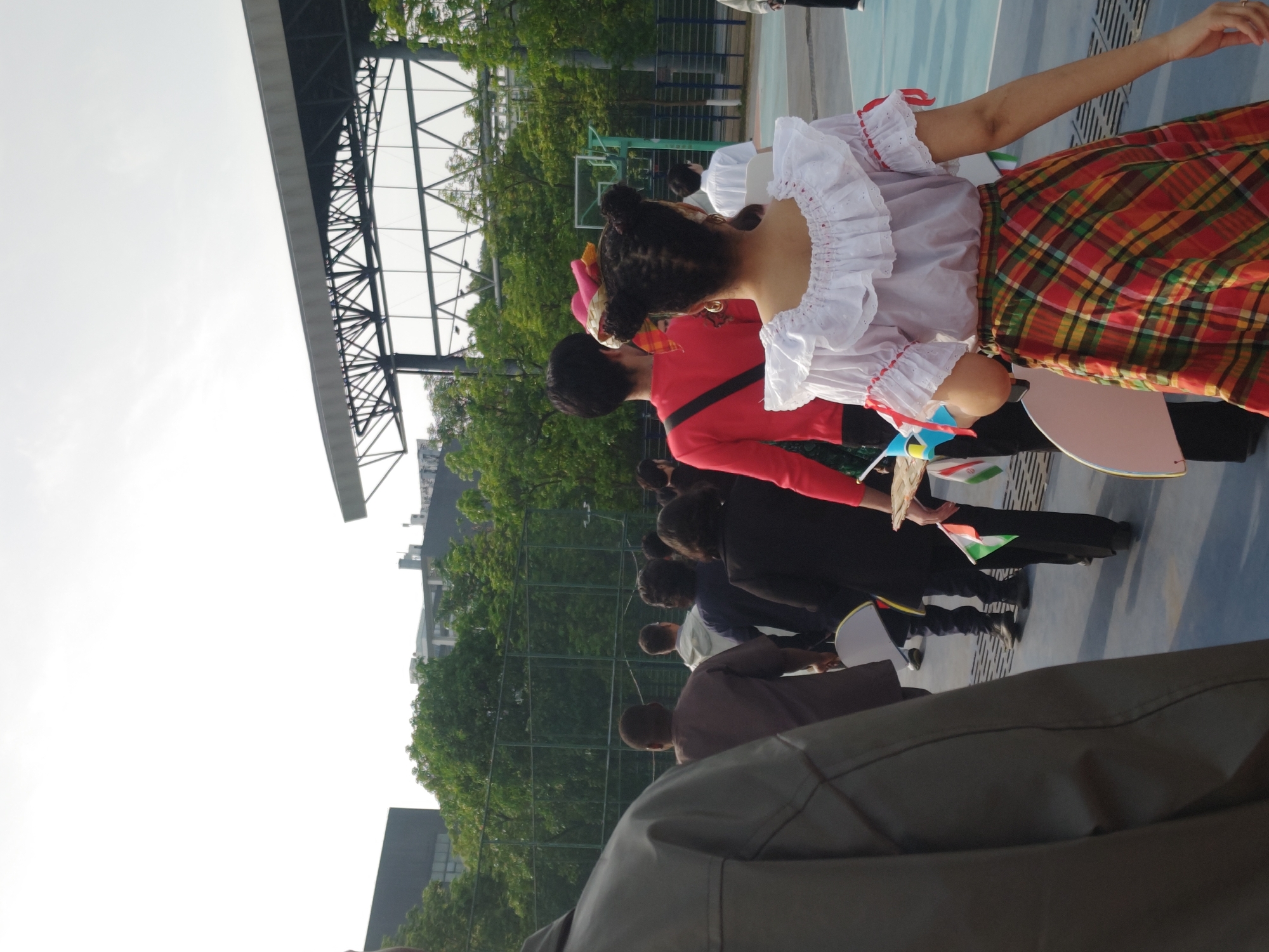 A group of people are participating in a parade or event on a light blue surface, likely a sports court, in what appears to be Chongqing High-tech Zone, China. A person at the front wears a white ruffled top and a plaid skirt, possibly a folk costume. The event is taking place outdoors under a metal structure with trees and buildings visible in the background. The group appears to be carrying flags.