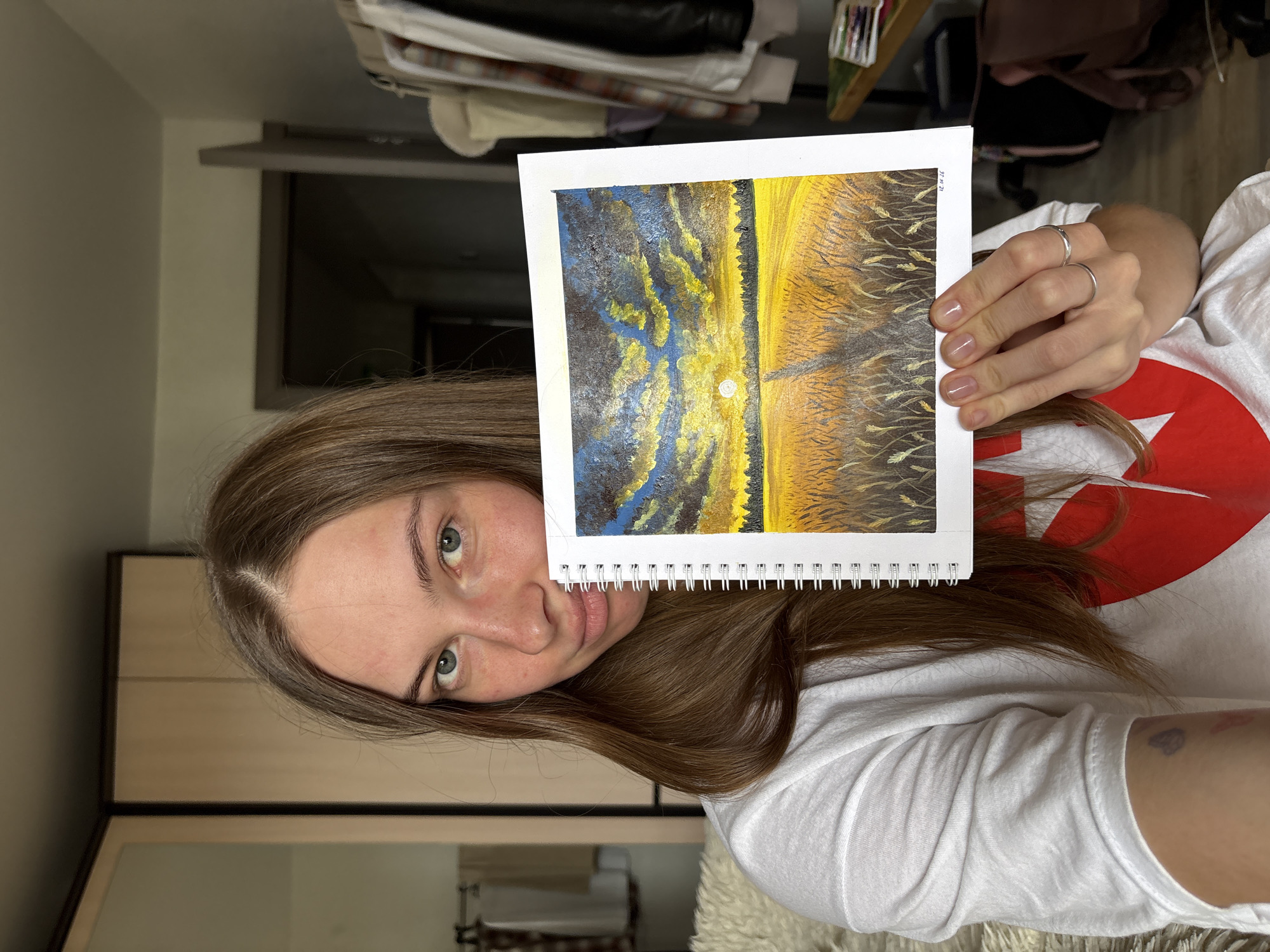 A young woman with long brown hair and blue eyes is holding up a painting of a sunset over a wheat field. She is wearing a white t-shirt with a red heart design. The background appears to be a bedroom with a closet and a rack of clothes. There is no indication of the city of Perm, Russia, in this image.