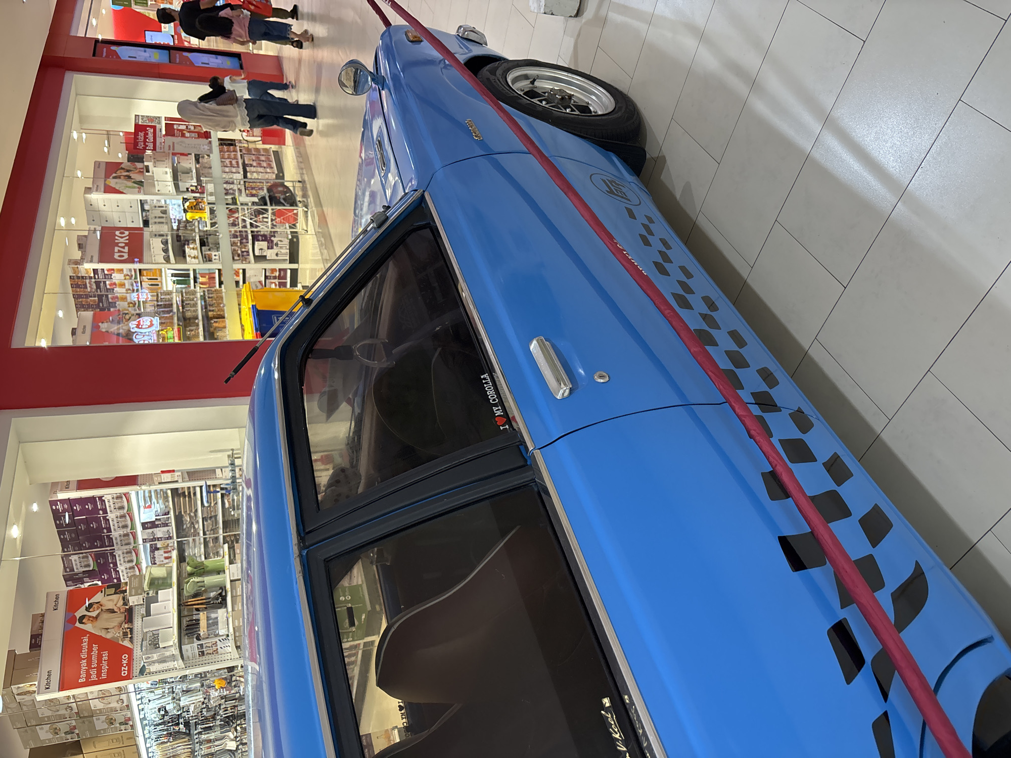 The image captures a blue car parked inside what appears to be a shopping center in Kuta, Indonesia. The car is positioned near a store with visible "Kitchen" and "az-ko" brand promotions. People are seen browsing the store, and the car is lined with a red rope. A sticker of "I love my Corolla" is pasted on the car door. "V-Kool" brand sticker is also pasted. A sticker with question "Apa kabar, M Bali Galeria?" meaning "How are you, M Bali Galleria?" in Indonesian is visible on the store glass window.