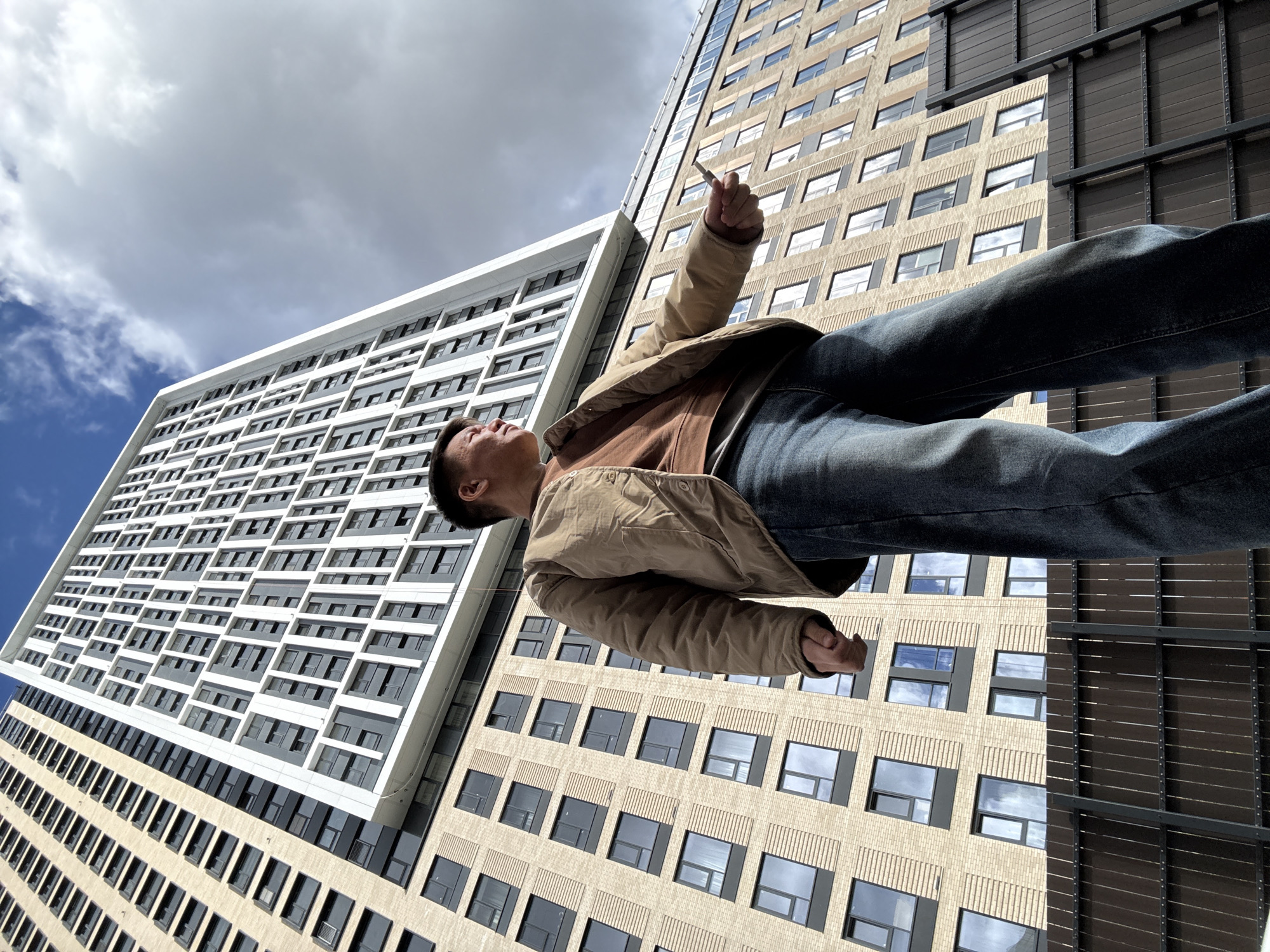 A man is seen from a low angle, seemingly lying on his back on the ground, facing upwards. He is wearing a beige jacket over a brown shirt, and blue jeans. He holds a thin, white object (possibly a cigarette or similar item) in his right hand, extended upwards.
Behind him are two tall buildings. The building on the left has a white facade with a grid of recessed rectangular shapes, likely windows. The building on the right has a tan facade with more regular, rectangular windows, and a section with dark, horizontal slats or panels.
The sky is visible between the buildings, showing patches of blue sky interspersed with clouds.
There is no information to confirm the location as Khimki, Russia, or any specific location at all.