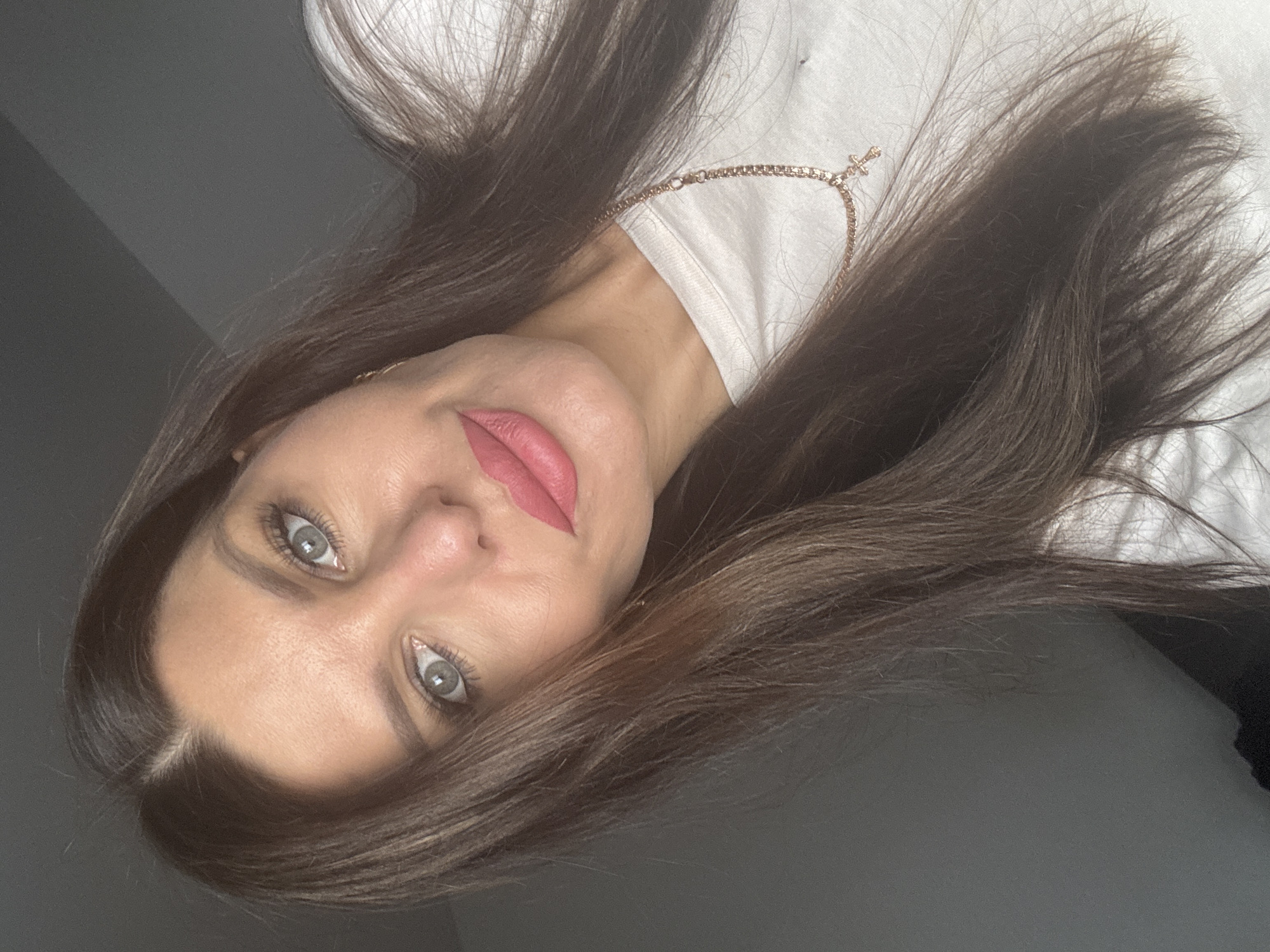 A woman is shown in a close-up portrait, looking towards the camera. She has long, dark brown hair, fair skin, and green eyes. Her lips are painted a vibrant pink, and she is wearing a white t-shirt with a rose gold necklace featuring a cross pendant. The background is a plain, gray wall, suggesting an indoor setting. There are no discernible urban or natural elements that would indicate a specific location in Chaikovsky, Russia.