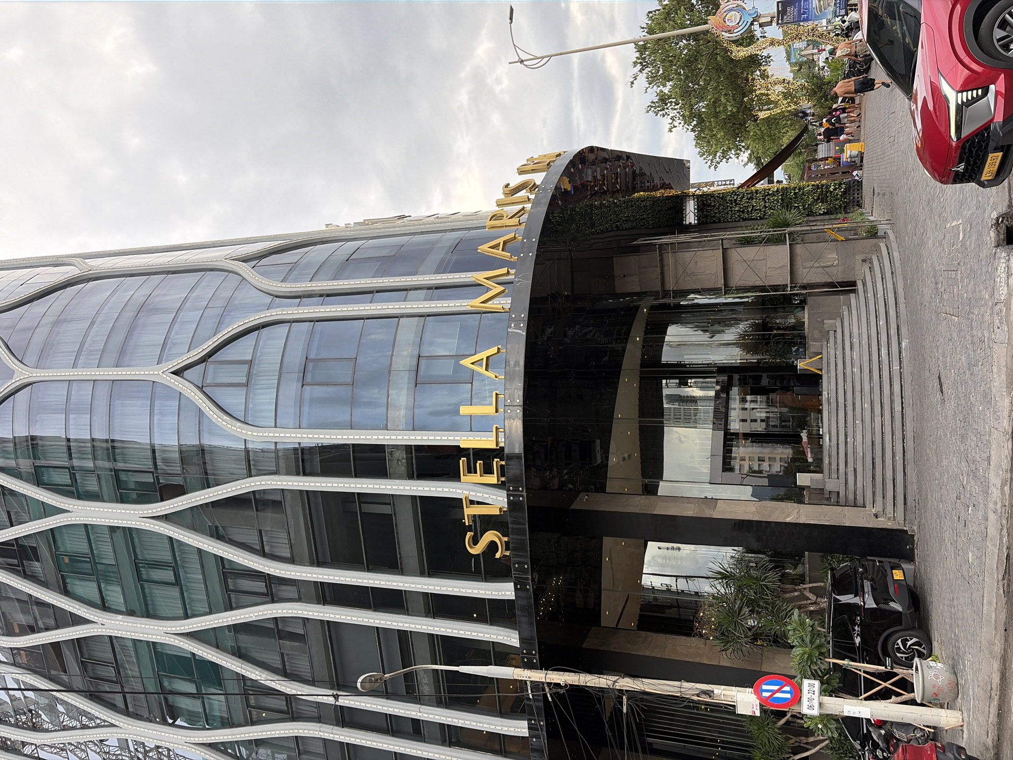 The image captures the Stella Maris Hotel, a tall, modern building in Đà Nẵng, Vietnam. The hotel features a curved facade clad in dark glass, with distinctive white, wave-like vertical accents. The hotel's name, "STELLA MARIS," is prominently displayed in large, golden, three-dimensional lettering above the main entrance.
At the base of the building, a wide, semicircular set of steps leads up to a glass-enclosed lobby. To the left of the entrance, a tall, slender lamppost stands next to a white pole displaying a circular traffic sign with red and blue markings and text indicating operating hours (06:00-22:00). A black SUV is parked to the left of the steps, with a portion of a red car visible in the lower right corner of the frame.
Beyond the hotel entrance, a paved walkway leads towards a street lined with trees and buildings. Several people are walking along this walkway, some appearing to be tourists. The sky is overcast, suggesting a cloudy day. The scene depicts a street-level view of a commercial area, likely a popular tourist destination given the hotel's presence and the pedestrian activity.