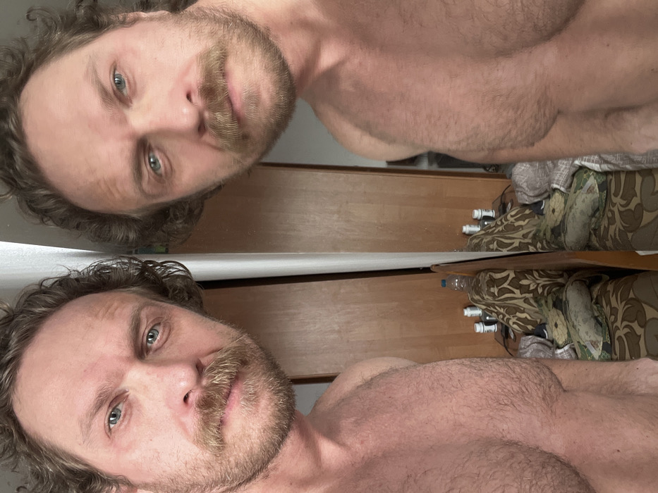 A male individual is depicted in a close-up, chest-up self-portrait taken in an indoor setting. He has medium-length curly brown hair, a full mustache, and a beard, with blue eyes. The man is shirtless, revealing a hairy chest and shoulders. He is positioned directly in front of a mirror, which reflects his image on the right side of the frame. The reflection shows a light brown wooden cabinet or wardrobe in the background. Below the reflected cabinet, a piece of furniture with patterned upholstery in brown and beige tones is visible. On this furniture, several white cylindrical containers and a clear plastic bottle with a blue cap are present. The scene indicates a private residential interior, located in Saint Petersburg, Russia.