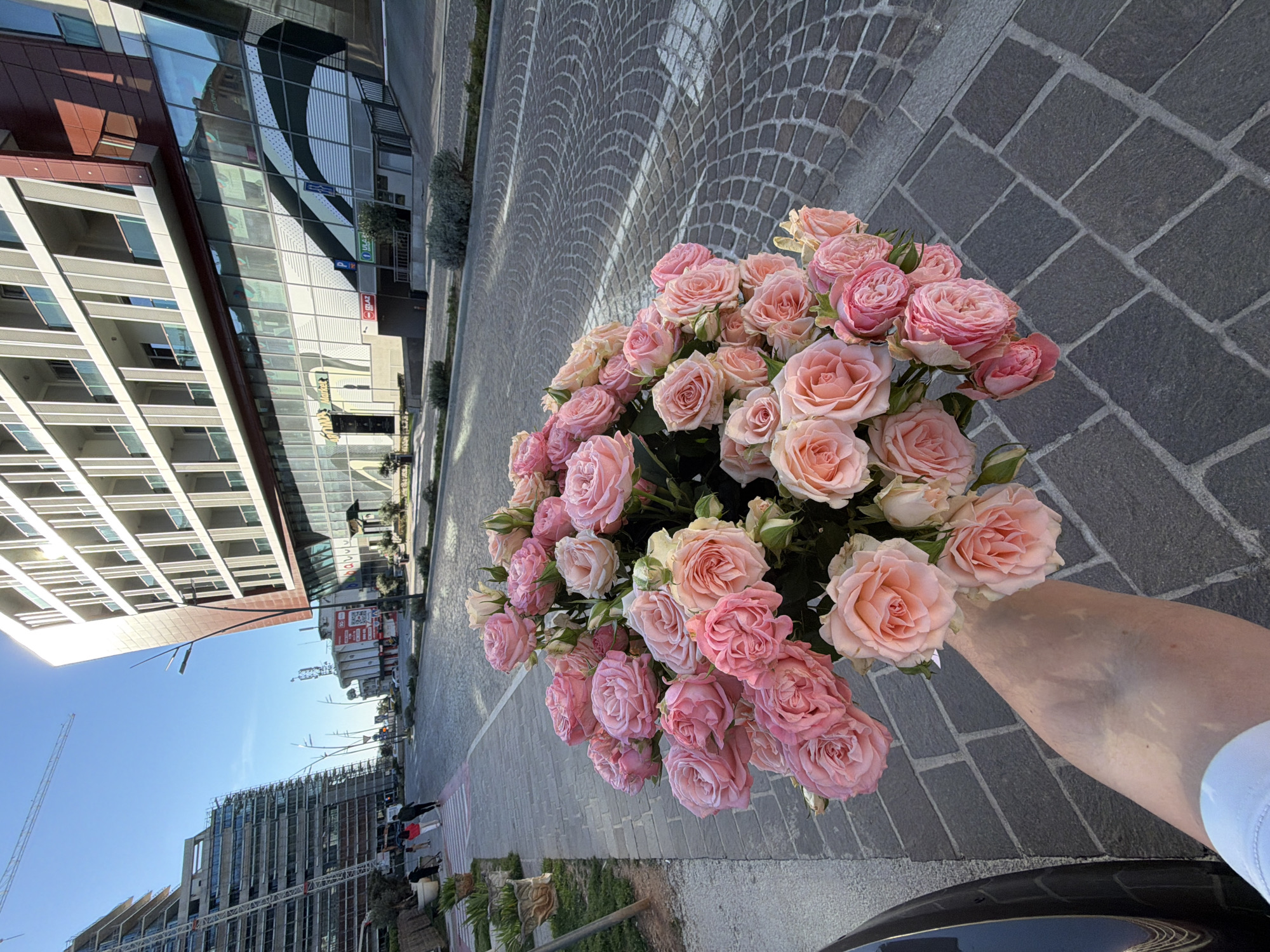 The image depicts a hand holding a bouquet of pink roses against an urban backdrop. The location is in Budva, Montenegro, as identifiable by the architectural style of buildings visible in the background. The foreground is dominated by the bouquet of roses, held by an arm extending into the frame from the bottom right. The background features a street paved with cobblestones, flanked by modern buildings with reflective glass facades. A building under construction with a crane is also visible. Several indistinct figures walk along the sidewalk. The sky is blue.