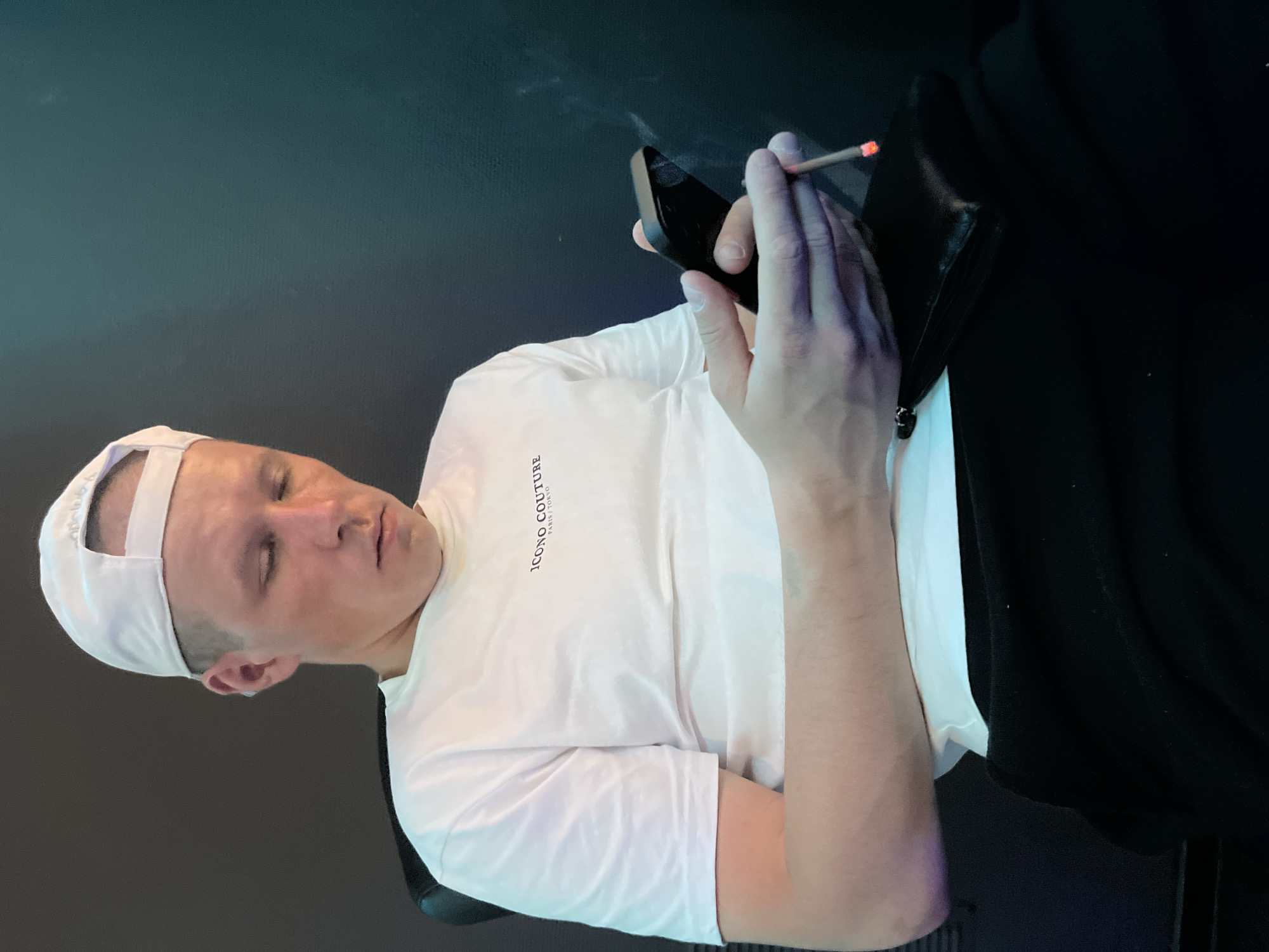 A male individual is seated indoors in an unknown city and region. He wears a white baseball cap, reversed, a white short-sleeved t-shirt with "ICONO COUTURE PARIS/TOKYO" printed in black on the chest, and black trousers. He has short-cropped hair and a small ear stud on his left ear. His head is tilted down, and his eyes are directed downward, appearing closed or focused on an object. He holds a black smartphone horizontally in his right hand. In his left hand, he holds a lit cigarette, from which a plume of smoke is visible ascending to the right. A black wallet or small pouch rests on his lap. He is seated in a dark chair, with a plain, dark teal or dark grey wall serving as the background. The lighting is subdued, illuminating the subject from the left side.