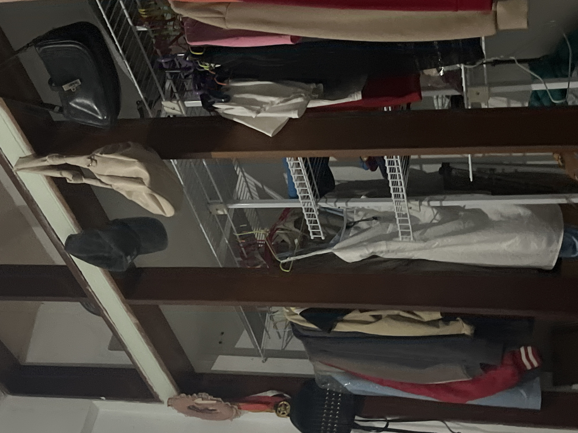 The image depicts an interior scene, possibly a closet or wardrobe area, with several items of clothing and accessories visible. In the upper section, two handbags are hanging from a wooden beam: a black structured handbag on the right and a lighter-colored, slouchy bag on the left. Below these, a black baseball cap is also hanging.
The middle and lower sections are filled with clothing on hangers, predominantly in muted or dark colors. There are several garments hanging on racks, including what appears to be a white dress or long top suspended in the center. The overall impression is one of a storage space for clothing, likely in a residential setting. The image does not provide any visual cues or context that would allow for identification of the location within Santa Ana, Venezuela.