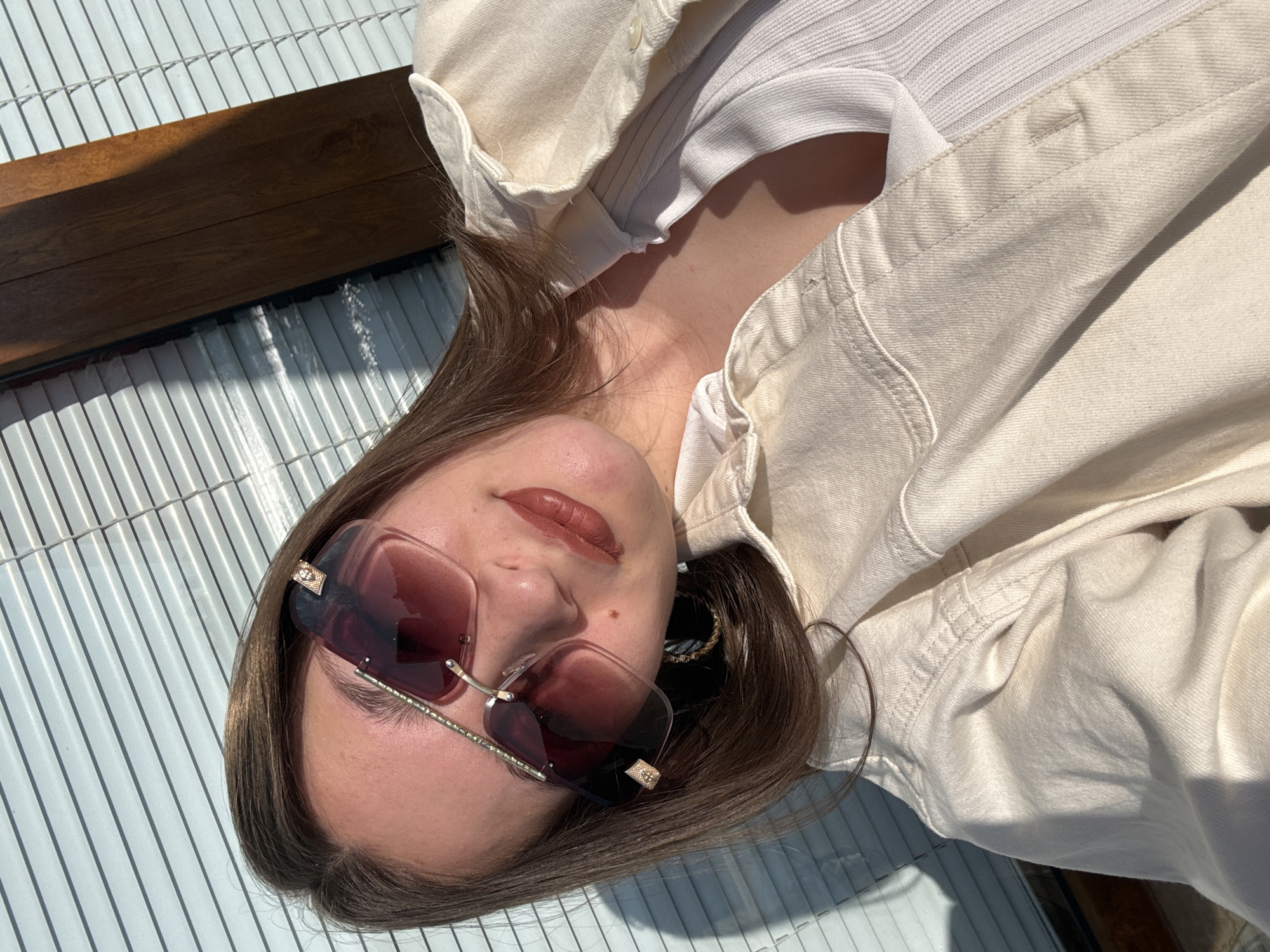 A woman is reclining outdoors, possibly on a tiled surface, wearing sunglasses and a light-colored denim jacket over a white top. She is facing the camera and appears to be relaxed. The background includes what looks like a striped surface or wall, and part of a wooden structure. The location in Sozopol, Bulgaria, is not evident from this image.