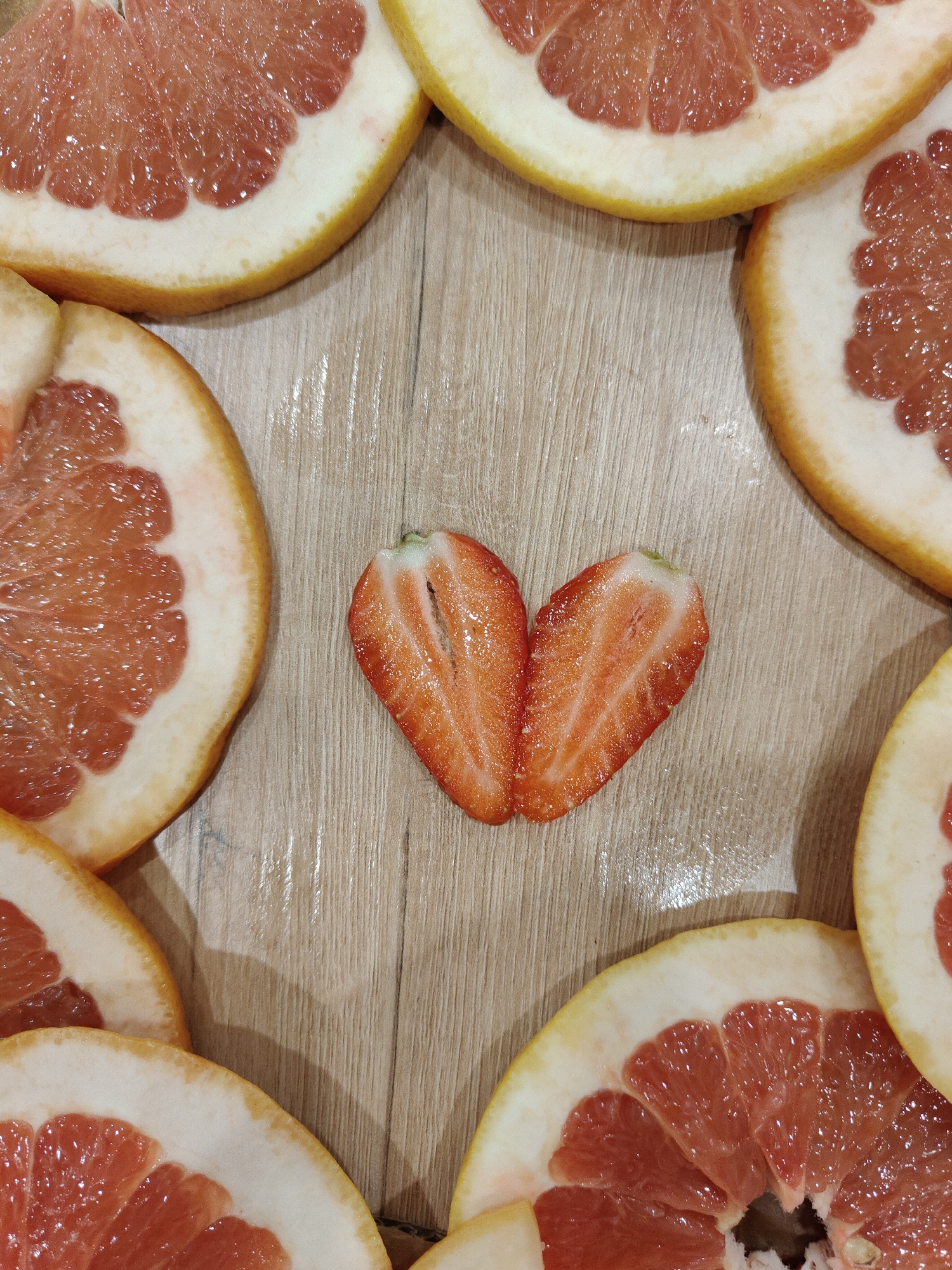 The image shows slices of pink grapefruit arranged in a loose frame around two halves of a strawberry, positioned to resemble a heart. The setting appears to be a wooden surface. The location is unknown.