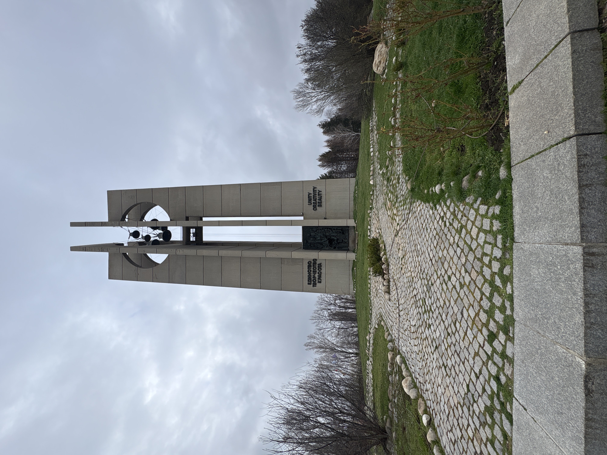 The image shows the Bells Monument in Sofia, Bulgaria. The monument consists of two tall, concrete pillars with an arch connecting them at the top. Hanging inside the arch are several bells. The inscription "UNITY CREATIVITY BEAUTY" is visible on the monument, along with "ЕДИНСТВО ТВОРЧЕСТВО КРАСОТА" in Bulgarian Cyrillic. A paved stone path leads towards the monument. The surrounding area includes grass, trees, and landscaping features. The sky is overcast.