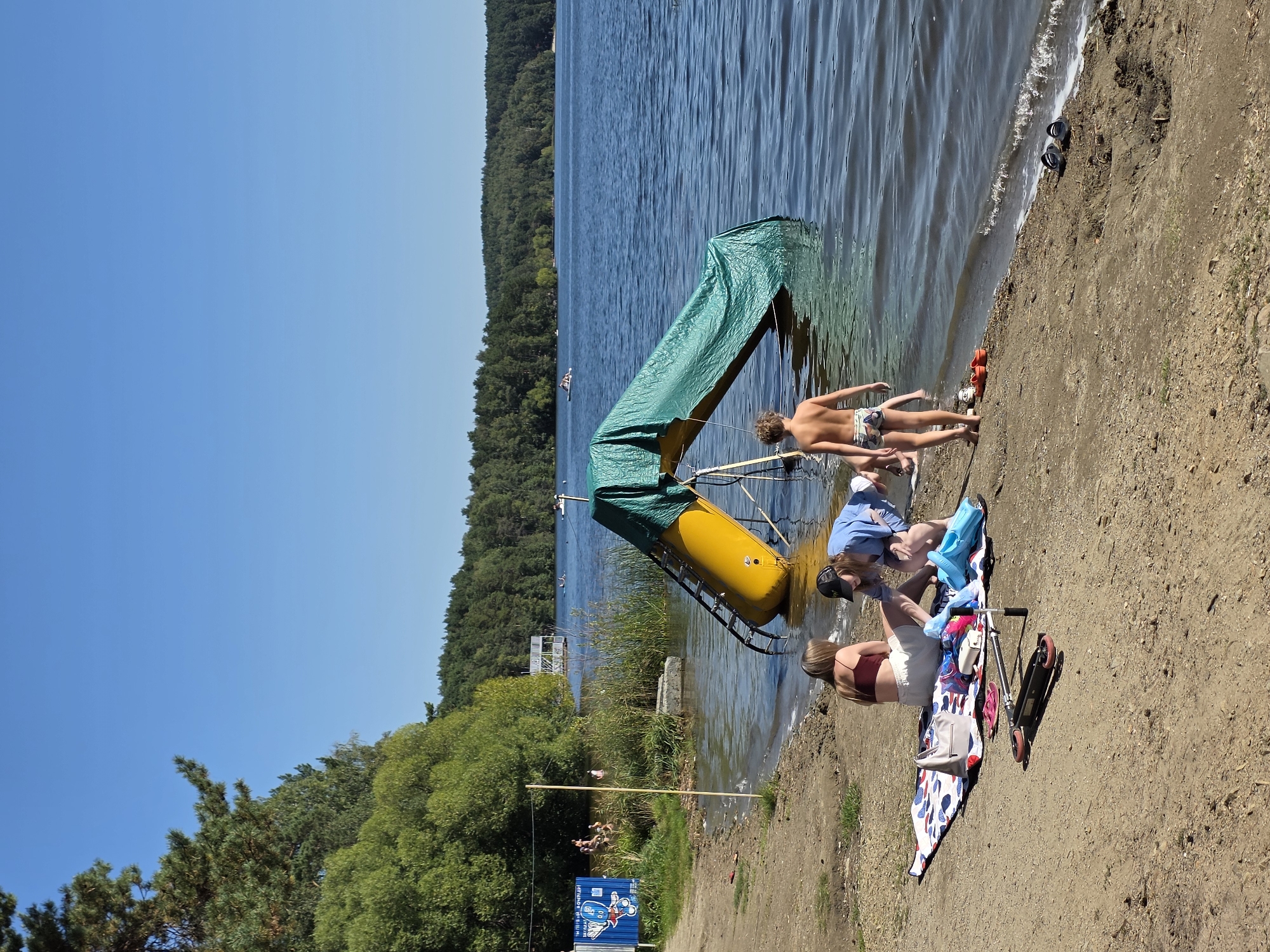 The image depicts a sunny day at a beach on the shore of Lake Chebarkul, Russia. The foreground shows a sandy beach with several people. Two women and a young boy are sitting on a blanket, with a scooter and beach toys nearby. The boy is wearing swim trunks and facing away from the camera, standing near the water's edge. In the water, a yellow slide covered with a green tarp extends from the shore into the lake. In the background, trees line the far shore of the lake, and several small boats are visible on the water. A wooden structure, possibly a pier or a viewing platform, is also present in the distance. A sign with cartoon graphics is posted on the left side of the image near the water. The sky is clear and blue.