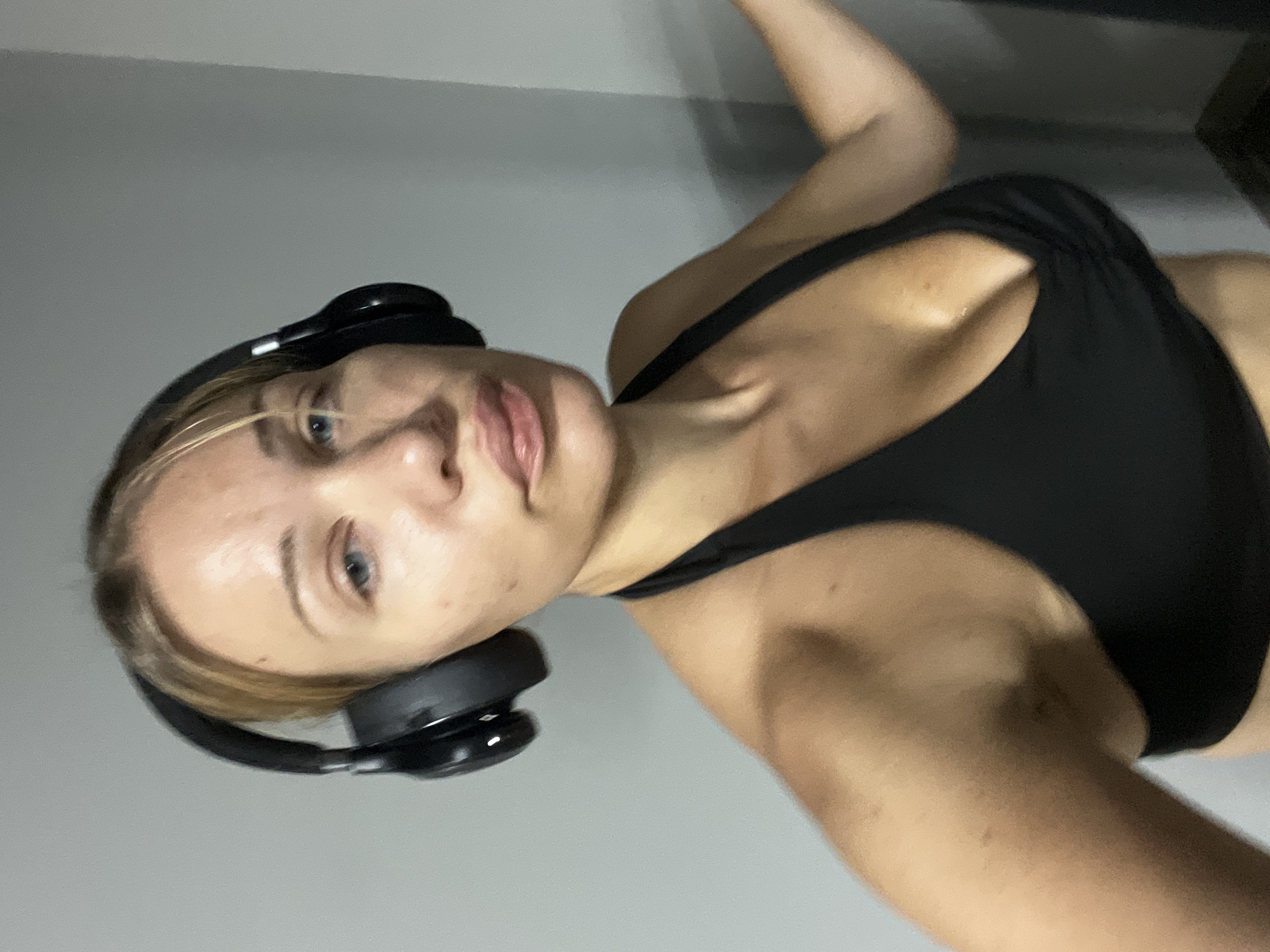 A woman with blonde hair and blue eyes is pictured from the chest up, wearing black over-ear headphones and a black sports bra. She is indoors, with a plain gray wall behind her. The lighting is somewhat dim, creating shadows on her skin. She appears to be looking at the camera. No location in Altötting, Germany is discernable from the image.