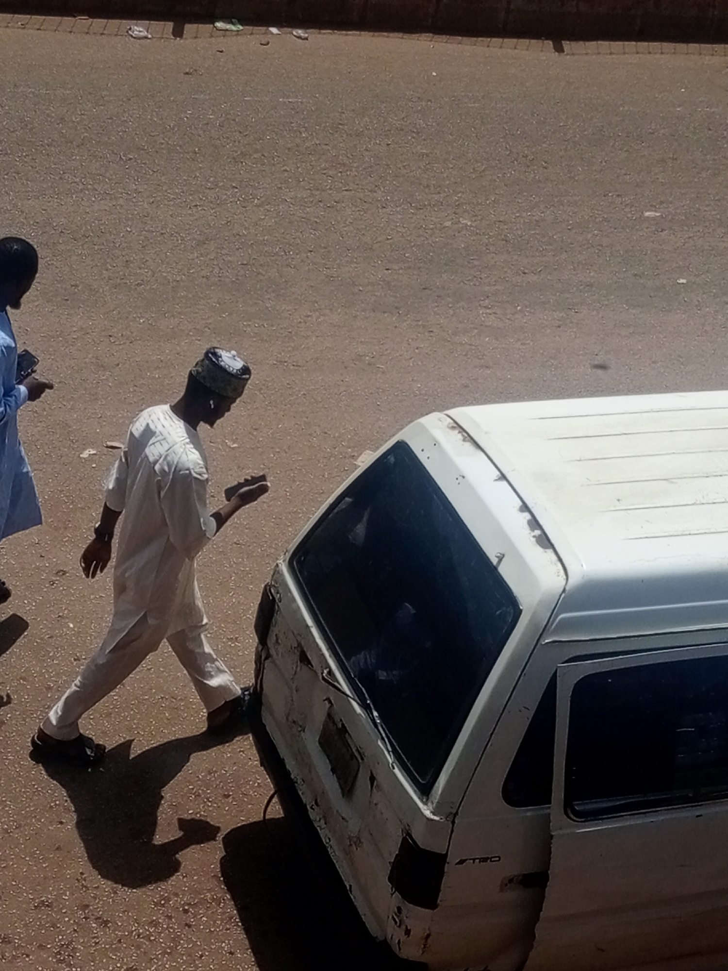 In Bukuru, Nigeria, an overhead shot captures a street scene with two individuals and a white van. The person on the left, partially visible, wears a light blue garment and holds a mobile device. To their right, a man in a white traditional outfit, including a patterned cap, walks with his right hand extended, holding a small rectangular object. His left hand is down by his side. He is walking past the rear of a white van, which appears to be aged and shows signs of wear, with some rust and damage visible on its exterior. The van has tinted windows. The ground is a textured, dusty asphalt surface. The sun casts distinct shadows, indicating a bright, daytime setting.