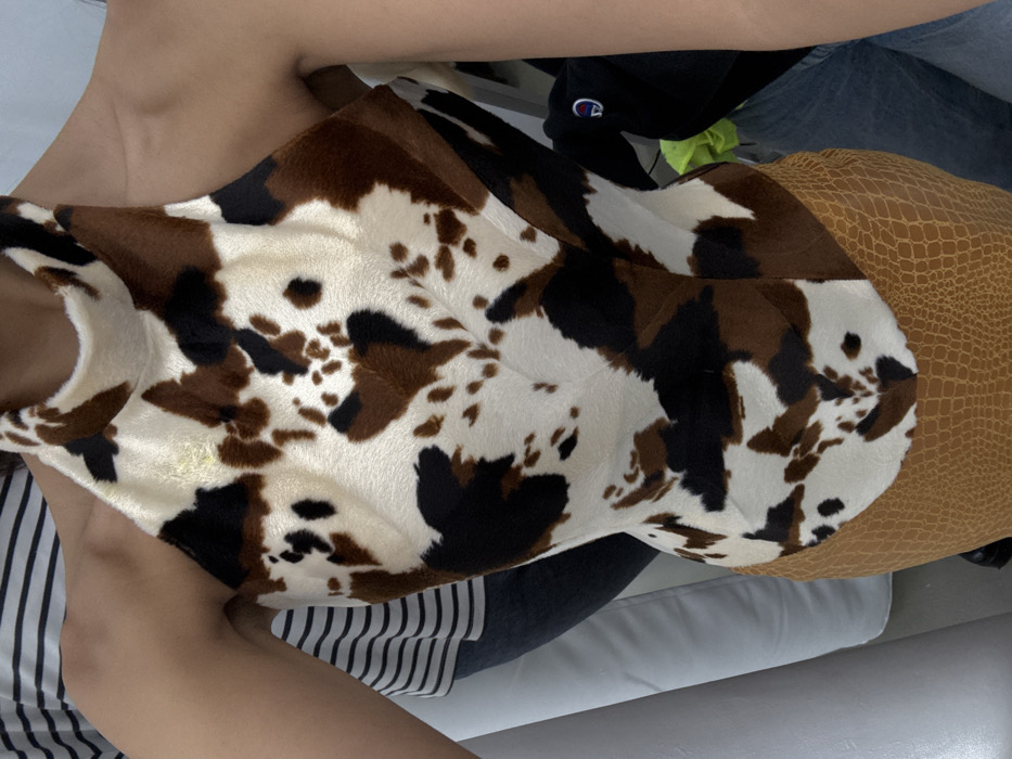 A person wearing a cow-print top and a brown, reptile-patterned skirt or shorts. The outfit is set against a background that includes a striped garment, a black baseball cap with a logo, and gray denim. The person is seated on a white surface.