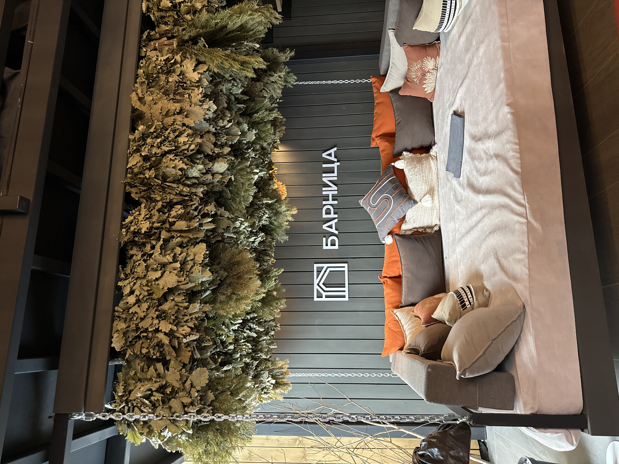 The image captures an interior space, likely in Vyakhtelevo, Russia, considering the text "БАРНИЦА" (BARNICA) which is a Russian word related to bathhouses. On the left, a wall is adorned with hanging bundles of what appear to be birch and evergreen branches, reminiscent of the traditional "venik" used in Russian bathhouses. The opposite side features a cushioned bench or daybed with multiple decorative pillows in shades of gray, brown, and orange. A dark-colored frame encloses the bench. The overall aesthetic is rustic and suggests a connection to nature and traditional Russian bathing practices. A silver chain hangs between the branches and the bench.