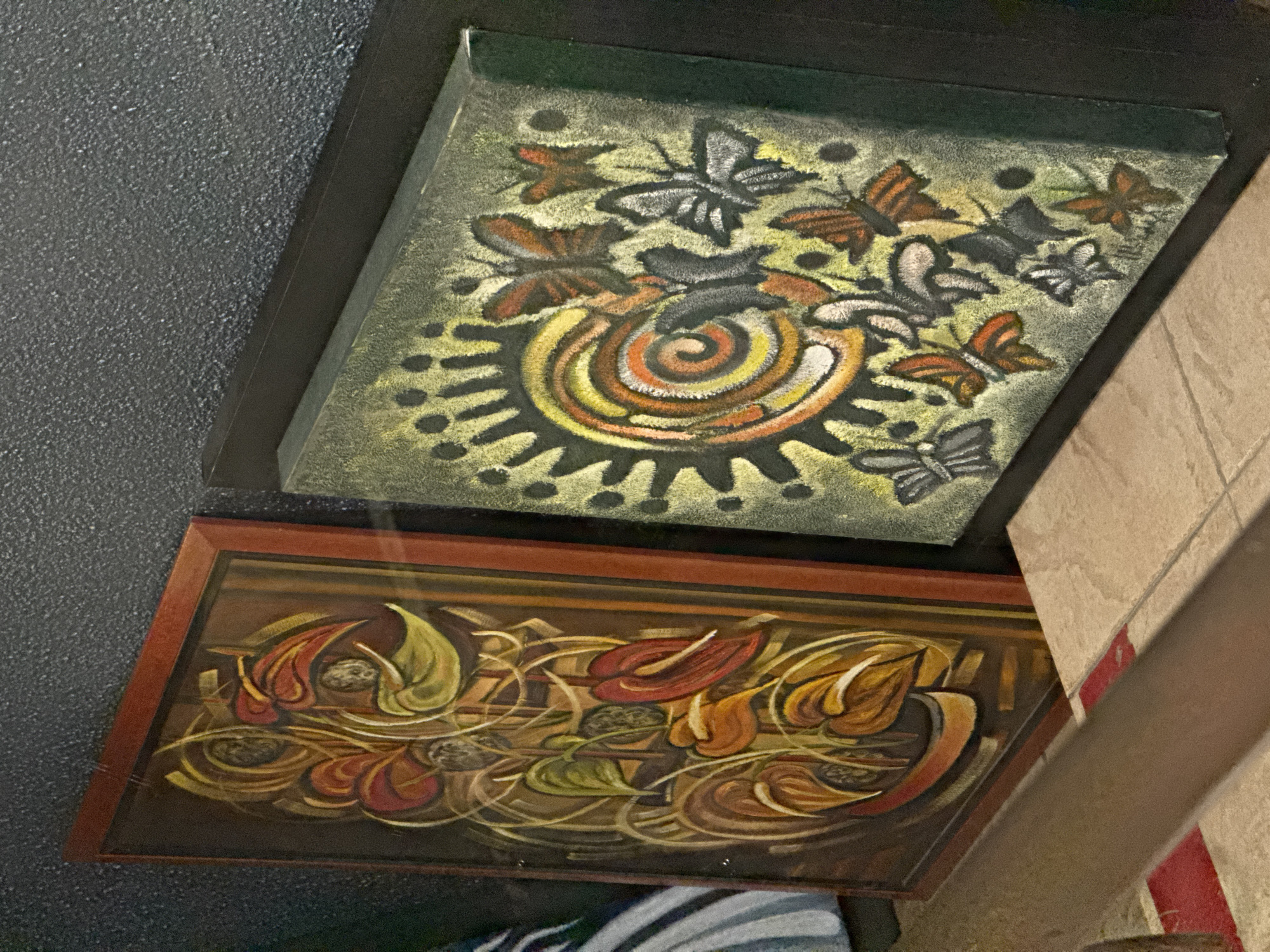 The image captures an angled view of two framed artworks displayed vertically on a dark, textured wall, likely within an interior space in Varna, Bulgaria.
The upper artwork, enclosed in a black frame, features a striking and tactile composition. It depicts numerous stylized butterflies in shades of orange, brown, gray, and white, arranged around a central sun-like or swirling motif. This central element combines vibrant yellows, oranges, and whites against a dark spiral, surrounded by dark, radiating dots. The background is a lighter green with yellow accents. The artwork appears to be made with a granular, sandy texture, giving it a unique dimensionality.
Below it, the second artwork, set in a dark wooden frame, showcases an abstract or highly stylized botanical design. It presents an intricate arrangement of organic forms resembling leaves or petals in rich tones of red, orange, yellow, and green. These elements are interconnected by flowing, light-colored tendrils against a warm, dark brown background. The style suggests movement and natural growth.
The wall behind the artworks is uniformly dark and features a coarse, pebbled texture. To the right of the frames, a section of a lighter-colored wall or architectural element is visible, featuring subtle vertical red stripe details. The lighting suggests an indoor setting, likely during the day or in a well-lit evening environment. No visible text is present on the artworks or their immediate surroundings.