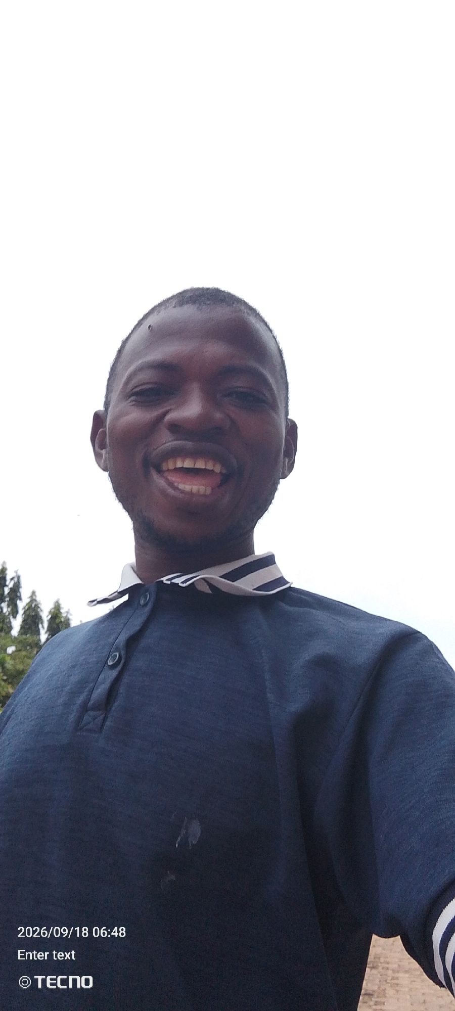 A close-up, low-angle shot shows a Black male with dark skin, short dark hair, and a light stubble on his chin and cheeks. He is smiling widely, with his mouth open and teeth showing, and his eyes are closed. He is wearing a dark blue polo shirt with a white and dark blue striped collar. The shirt has two buttons at the neck, one of which is buttoned. A small dark mark is visible on the shirt's chest. The background is a plain, overcast sky. In the lower left corner of the image, text reads "2026/09/18 06:48 Enter text © TECNO".