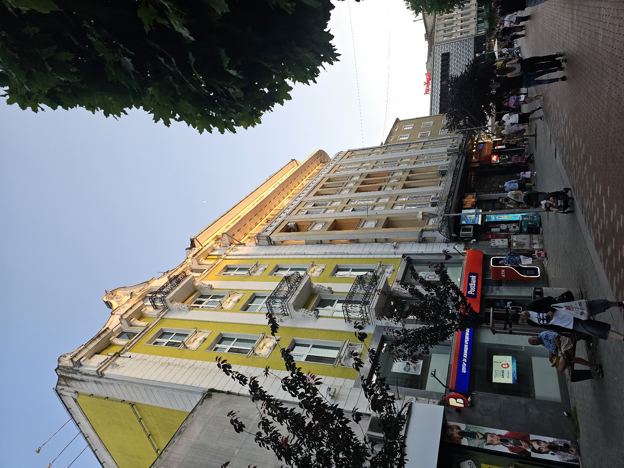 The image depicts a street scene in a city, with a focus on the architecture of the buildings and the pedestrian activity on the sidewalk. The buildings lining the street are multi-story, with some featuring ornate facades and balconies. The building on the left is painted a bright yellow and white, while the building to its right is a lighter beige color with many windows. A large tree with dark leaves partially obstructs the view of the buildings on the left.
On the sidewalk, people are walking in both directions, and some are sitting on a bench in front of a bank. The bank has prominent signage for "Postbank". Further down the street, more shops and businesses are visible, with some displaying illuminated signs. The sky above is clear and light blue, suggesting it is either early morning or late afternoon. The ground is paved with bricks. There are streetlights and some overhead wires visible. The overall atmosphere is that of a typical urban street with commercial activity and pedestrian flow.