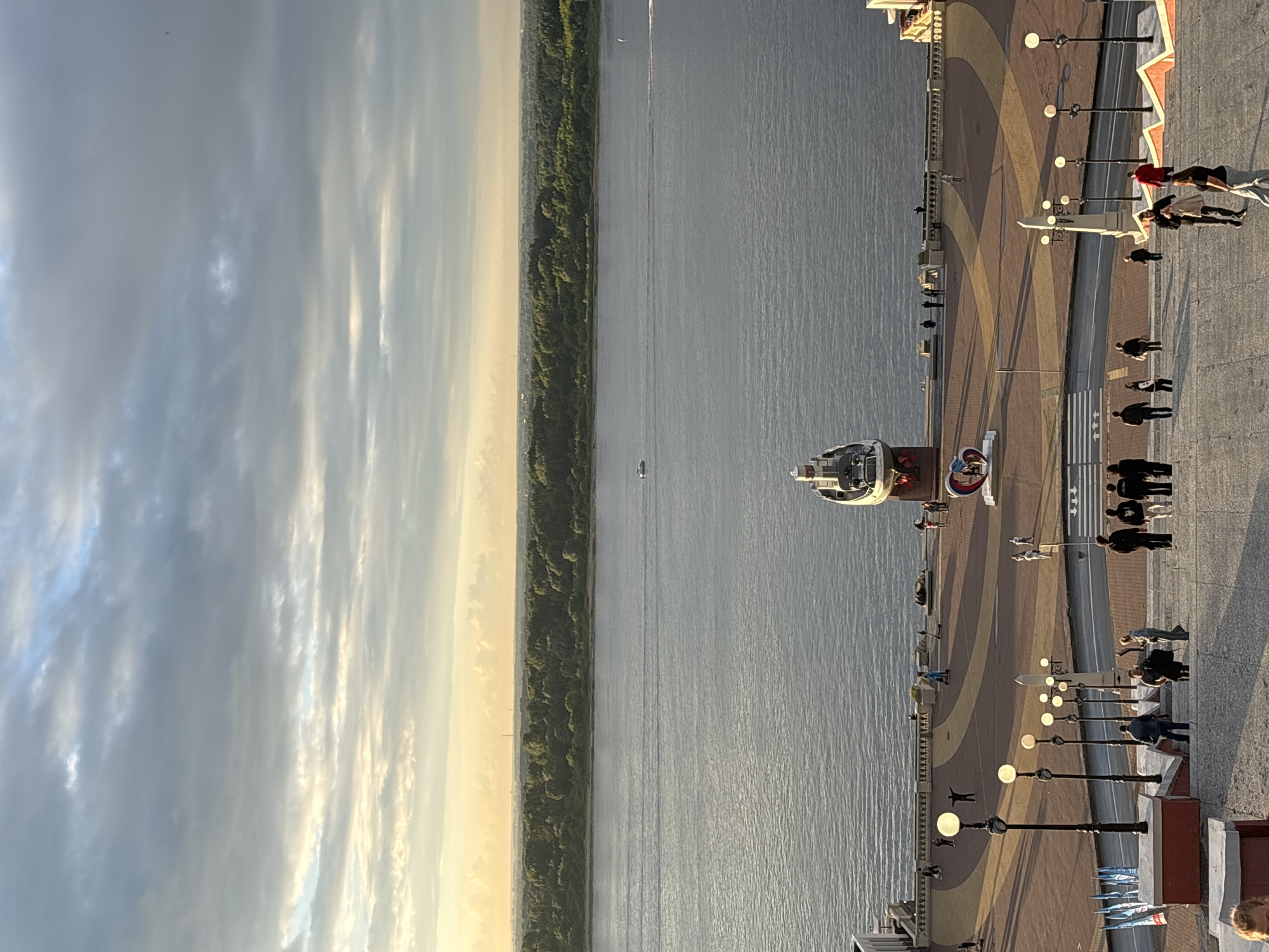 The image depicts a wide river with a treelined bank on the opposite side, under a sky filled with dramatic clouds and streaks of golden light from a setting or rising sun. In the foreground, a wide paved area with decorative patterns leads down to the river embankment. Several people are visible walking on this paved area and on stairs leading down from the viewer's perspective. A large ship is docked at the embankment, and a heart-shaped sculpture is positioned in the middle of the paved area. The scene is located in Nizhny Novgorod, Russia, likely at the Nizhny Novgorod Kremlin or a nearby viewpoint overlooking the Volga River. The overall atmosphere is one of a serene late afternoon or early morning, with a mix of natural beauty and urban elements.