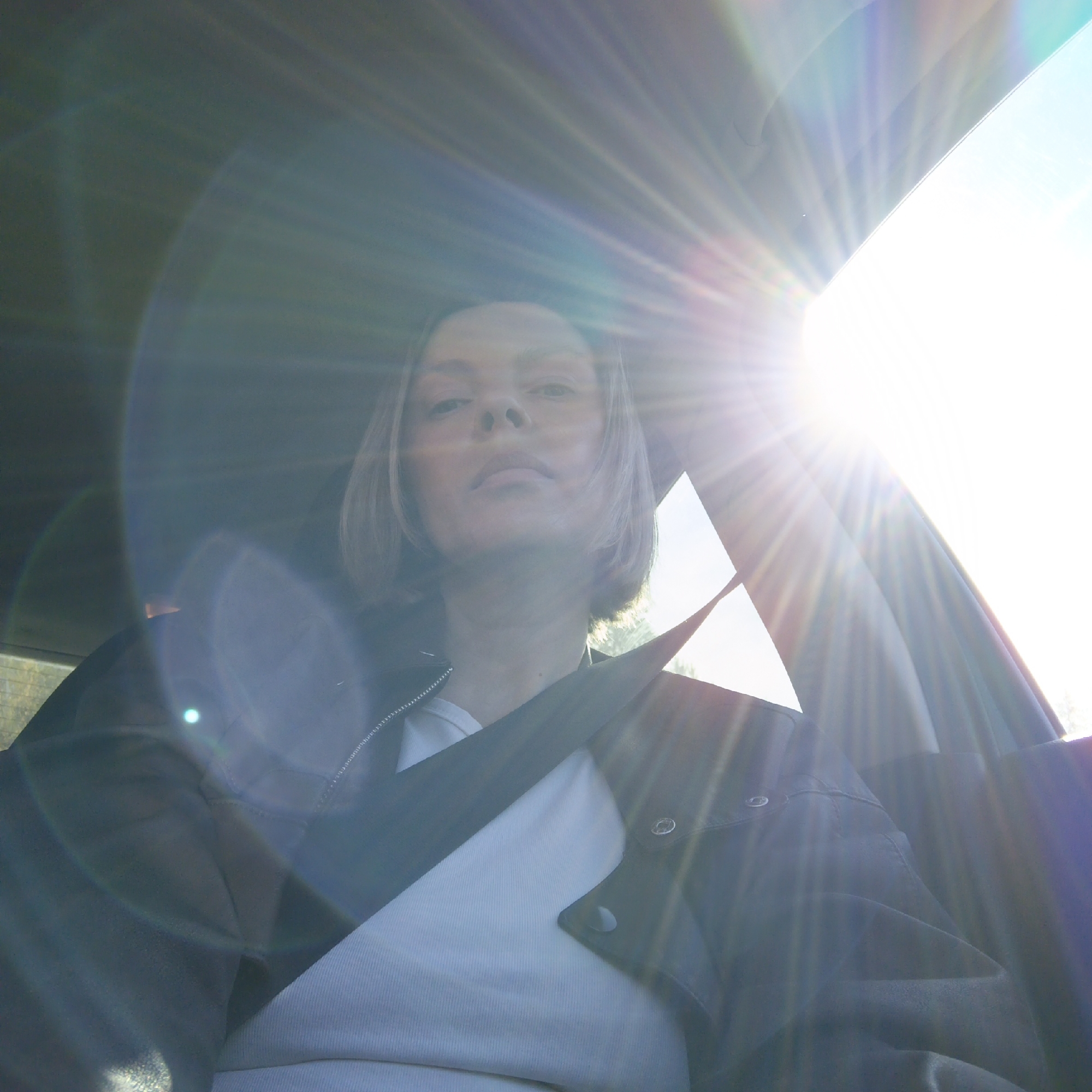 A woman with short blonde hair is sitting in a car, wearing a black leather jacket over a white top and a seatbelt. The sun is shining brightly through the car window, causing lens flare. The location is unknown and cannot be determined from the image.