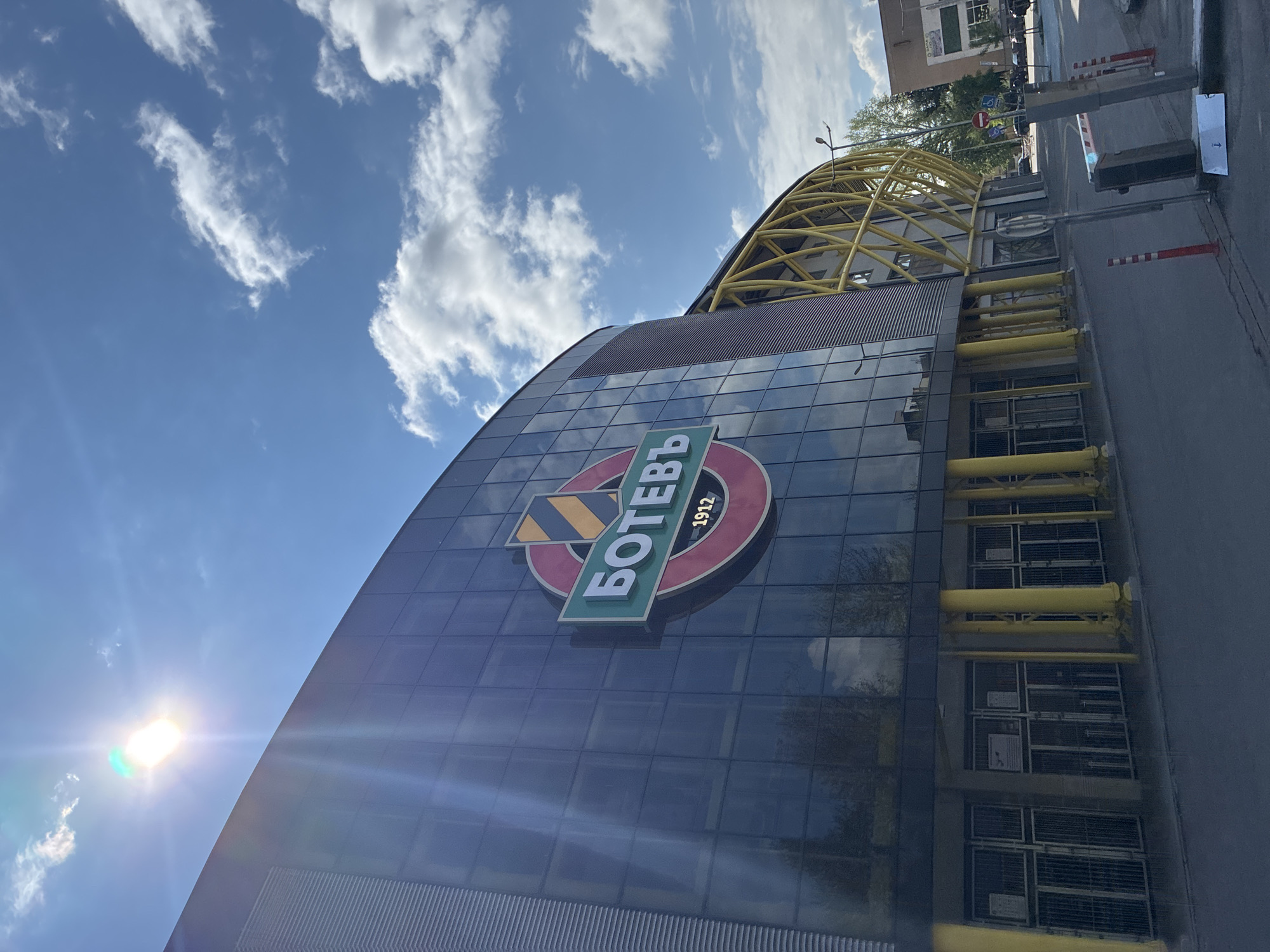 The image captures the exterior of the Botev Plovdiv stadium in Plovdiv, Bulgaria, under a partly cloudy sky. The modern, curved facade is primarily constructed of glass panels that reflect the sky. A prominent logo featuring the word "Ботев" in Cyrillic script and the year "1912" is affixed to the building's surface. A yellow framework is visible above the main structure. To the right of the building is a street or parking area with a barrier and bollards. The sun is visible in the lower-left corner of the frame.