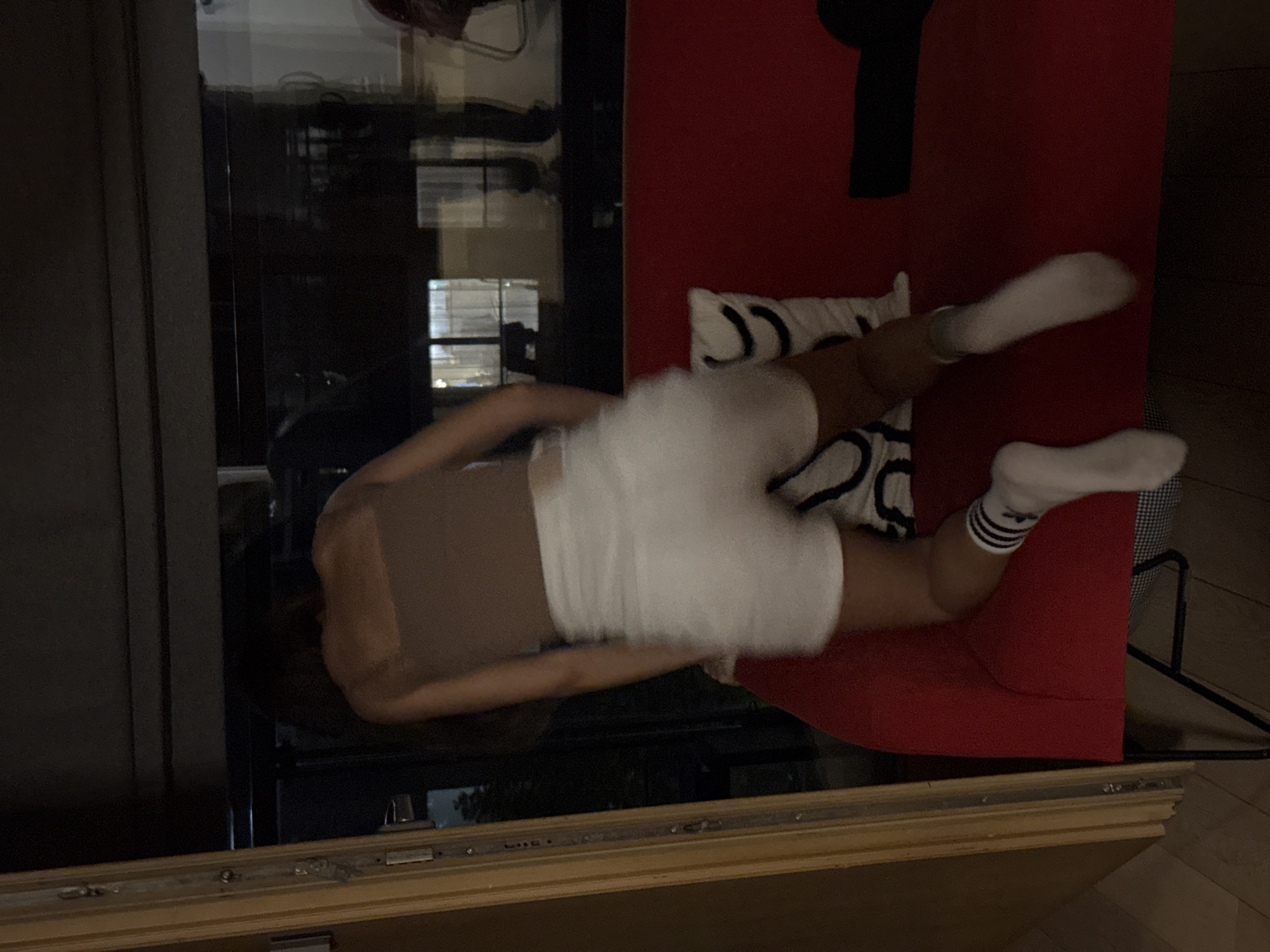 A person wearing a brown tank top and white shorts is lying on a red sofa with a white pillow. The person is also wearing white socks, one with black stripes. The sofa is next to a glass window or door. The environment appears to be indoors. The location is unknown.