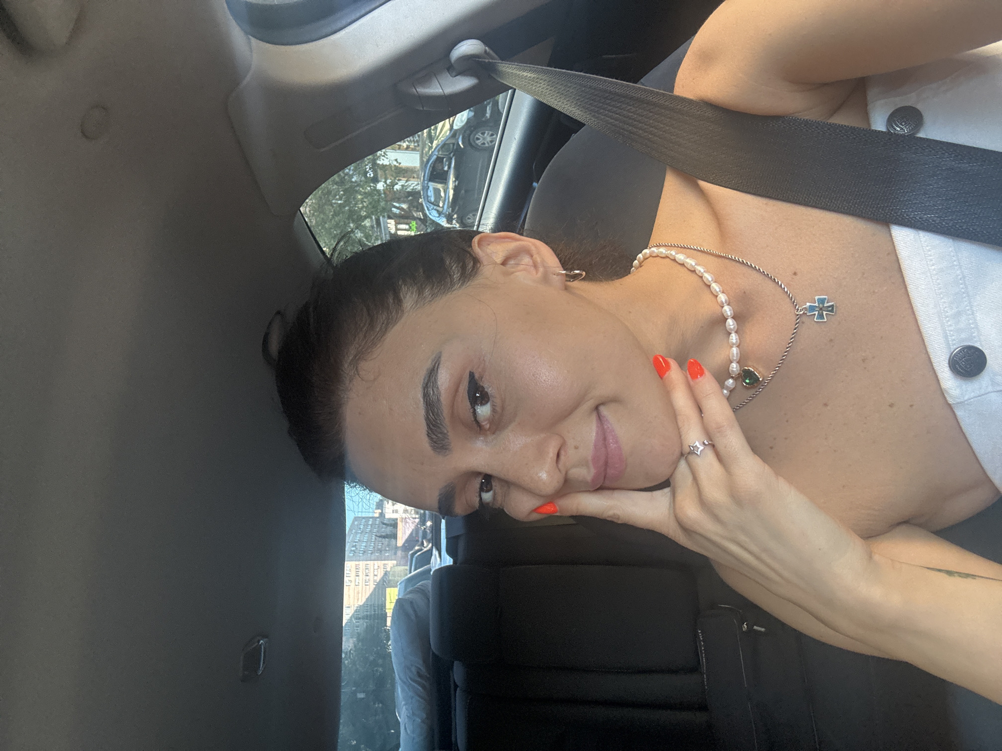 A woman is sitting in the passenger seat of a car, looking at the camera. She has dark hair pulled back in a bun and is wearing makeup, including winged eyeliner and lip gloss. She has bright orange nail polish on her fingers, which she is resting on her cheek. She is wearing two necklaces, one with pearls and a green heart-shaped pendant, and another with a cross pendant. The woman is wearing a white strapless top. The interior of the car is visible, including the back seat and a seatbelt. Outside the car, trees and buildings can be seen, suggesting an urban environment. The image appears to be taken during the daytime.