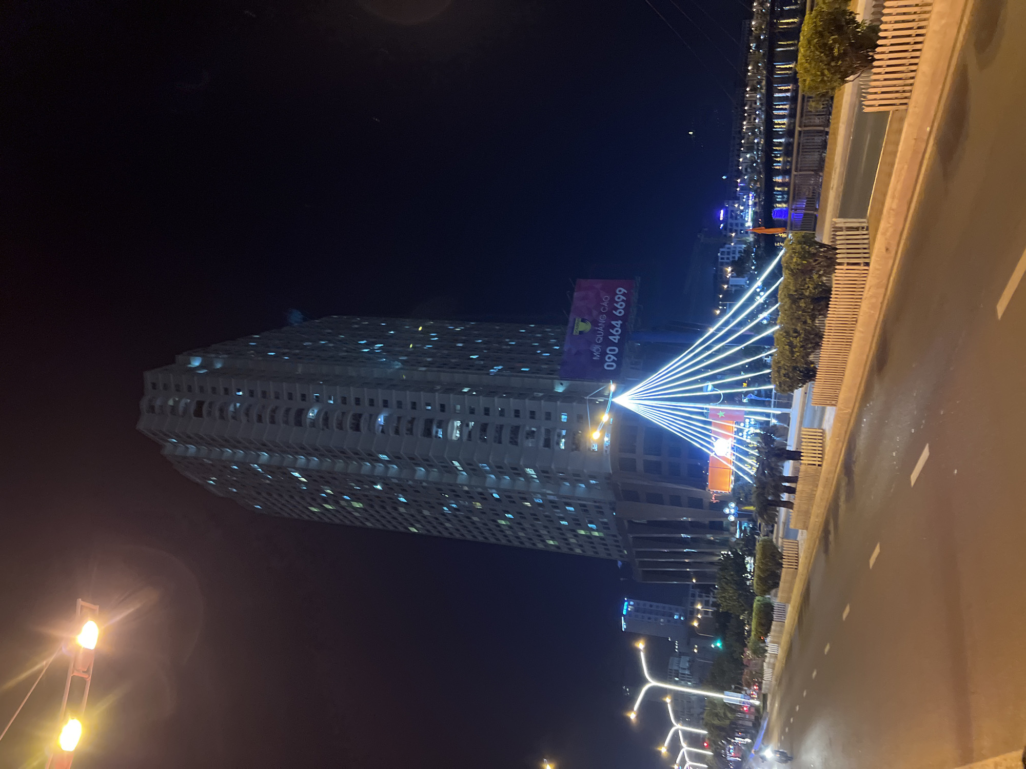 This is a night-time, eye-level, wide shot of a street in Nha Trang, Vietnam, featuring a tall apartment building, a bridge with decorative lighting, and a busy road. The apartment building is illuminated with numerous lights, indicating residential occupancy. The bridge is adorned with a conical arrangement of white string lights, creating a visually striking centerpiece. The road is paved and marked with white lane dividers, with a few streetlights visible, casting a warm glow. A motorcycle can be seen on the left side of the road, suggesting vehicular traffic. On the right side of the image, a body of water is visible, possibly a river or the sea, with buildings lining its opposite bank. A billboard advertising "MỞ QUẢNG CÁO" and a phone number is also present. The overall scene depicts a vibrant urban environment at night.