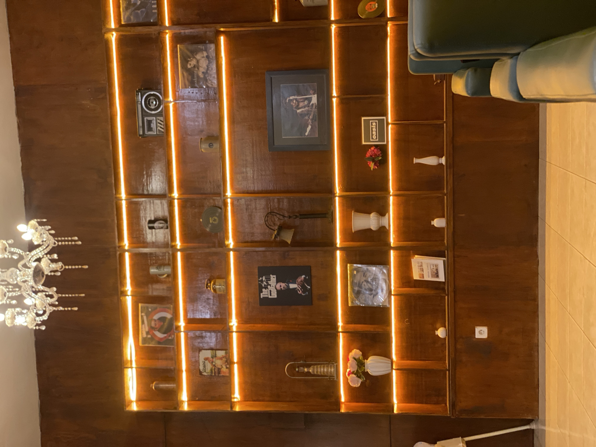 The image depicts a wall-mounted shelving unit with numerous items displayed. The shelves are illuminated by orange LED strip lights. On the shelves, there are vinyl records, framed pictures (including what appears to be a "The Godfather" poster, a framed image of a musician, and an "Oasis" album cover), decorative vases, mugs, a vintage camera, and other miscellaneous decorative objects. A crystal chandelier hangs from the ceiling above the shelves. In the foreground, the corner of a green armchair is visible, suggesting this is an indoor seating area. The overall aesthetic suggests a retro or vintage-themed establishment. The scene is likely a cafe or a lounge area in Banjarbaru, Indonesia, given the reference to the city.