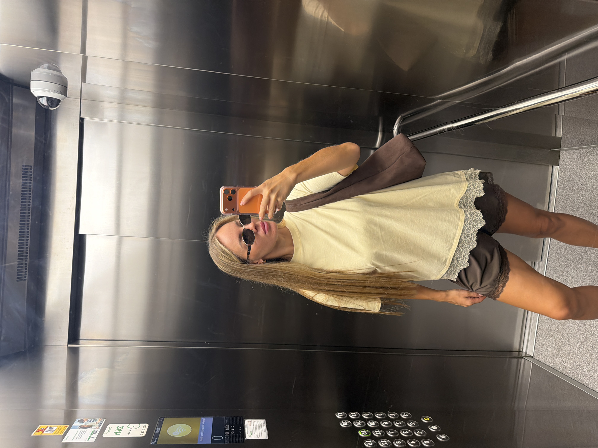 A young woman with blonde hair, wearing sunglasses, a light yellow t-shirt with lace trim, and dark shorts, takes a mirror selfie in an elevator. She is holding an orange-cased smartphone in her right hand. A brown bag is slung over her left shoulder. The elevator has stainless steel walls and a mirrored surface, reflecting the woman and the interior of the elevator car. A security camera is visible mounted on the ceiling in the upper left corner. The elevator buttons are visible on the lower right side of the frame, and there are some stickers on the wall in the lower left. The scene appears to be indoors. The overall mood is casual and confident.