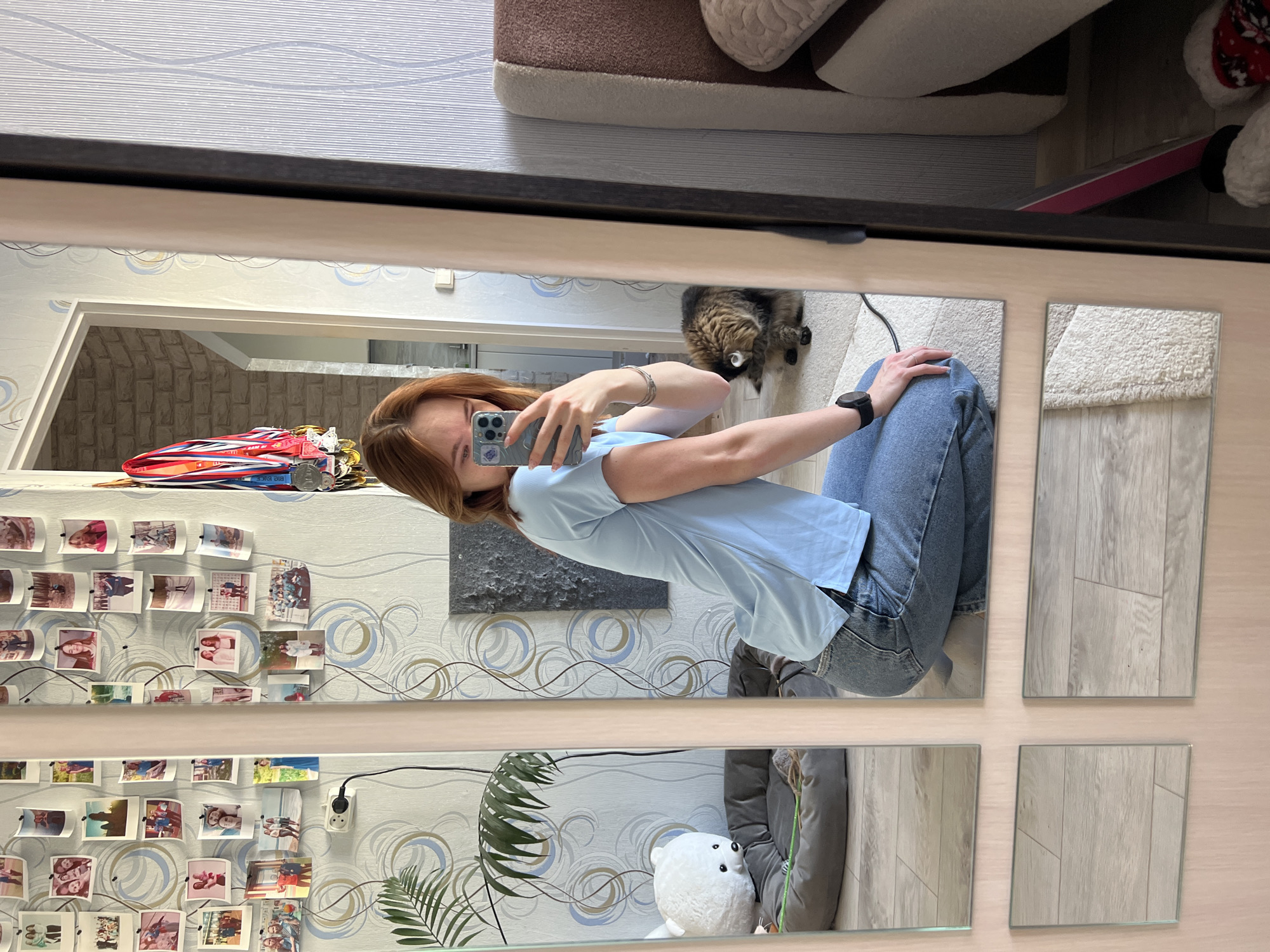 A woman is reflected in a fragmented mirror, holding a smartphone to take a picture of herself. She is wearing a light blue t-shirt and blue jeans, with a black wristwatch on her left wrist. The mirror is surrounded by a dark frame and reflects a room with patterned wallpaper, adorned with multiple small photographs and medals. A cat is visible in the reflection, near the top right. A stuffed polar bear is visible near the bottom of the mirror. The location is Chaikovsky, Russia.