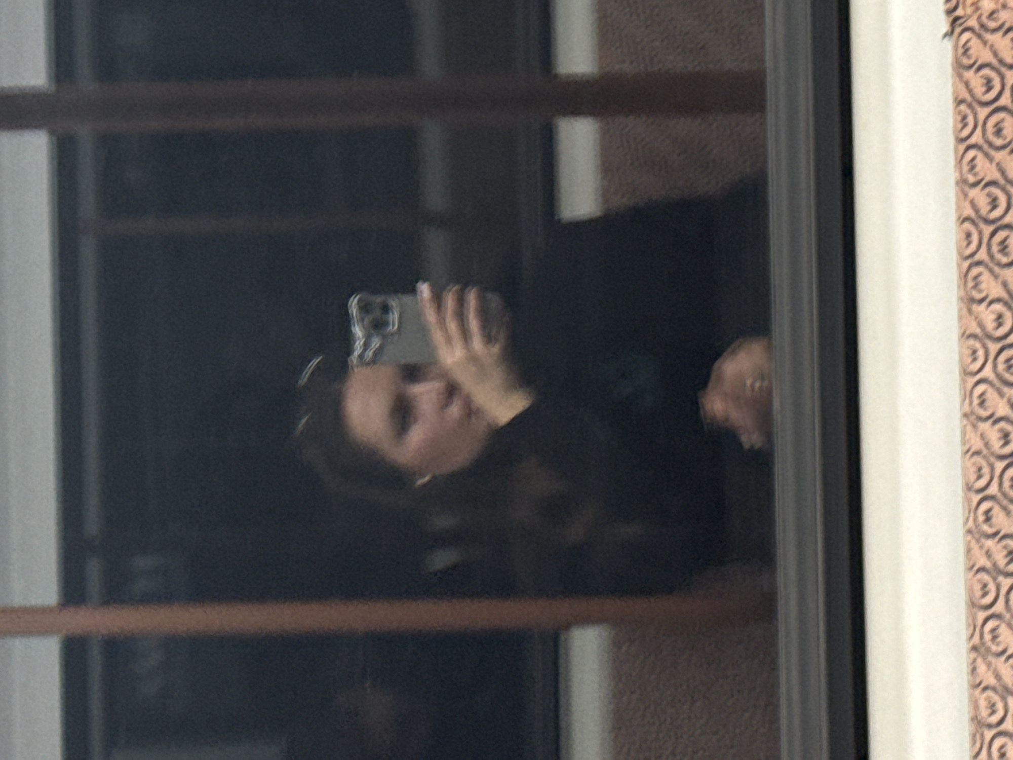 A person is reflected in a window while holding a phone. The window has a dark frame and is set against a wall with patterned wallpaper. The reflection also shows parts of the window frame and the outside environment is obscured. The location, Moscow, Russia, cannot be determined from this image alone.
