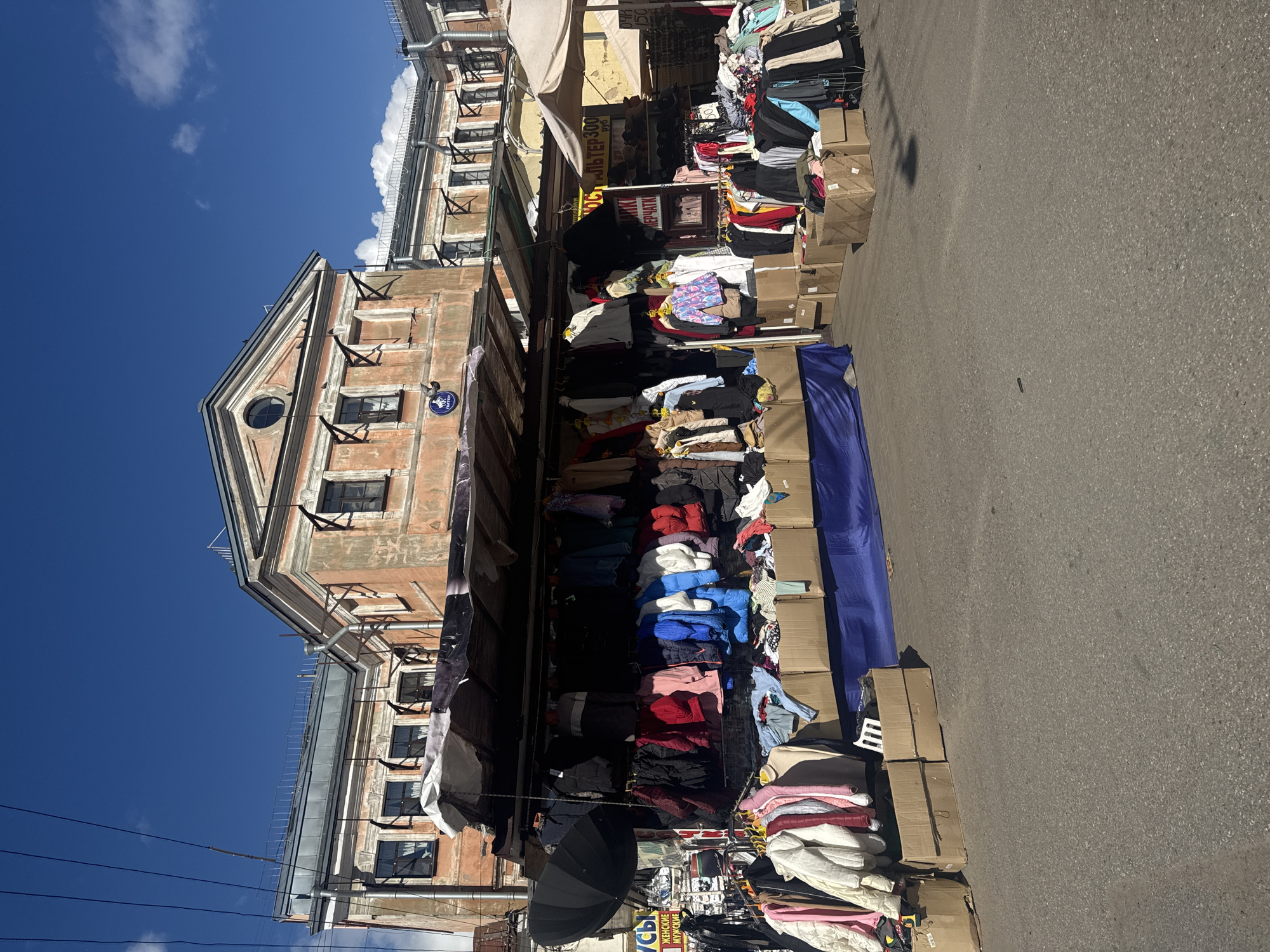 A street market stall is set up in front of an old, weathered building in Saint Petersburg, Russia. The stall displays a variety of clothing, including jackets, sweaters, and blouses, hung on racks and piled in cardboard boxes. A large black umbrella is partially visible on the left side of the stall. The building behind the stall has a distinct architectural style with arched windows and decorative trim. The sky is clear and blue with a few white clouds. The ground is paved with asphalt. There are no people visible in the image.