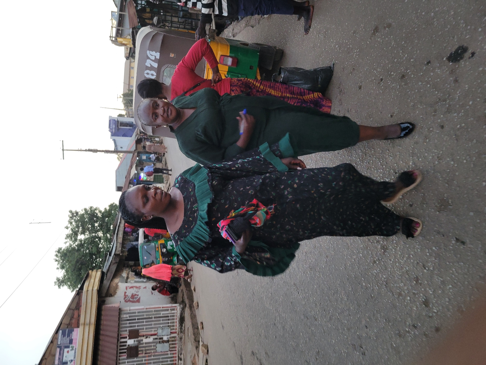 This image, rotated 90 degrees counter-clockwise, depicts a street scene in what appears to be an urban or semi-urban area, consistent with Jos, Nigeria. The lighting suggests it is daytime, possibly late afternoon or an overcast day, with soft, even illumination.
In the foreground, two women are prominently featured, looking towards the camera. The woman on the left (closer to the camera) wears a long, dark dress adorned with a floral pattern and pleated green details around the neckline and sleeves. Her hair is styled in braids. She holds a small, colorful bundle of fabric in one hand and a dark object, possibly a phone, in the other. Her feet are visible in open-toed sandals with red nail polish. The woman on the right wears a plain dark green long dress and has short, grey hair. She holds a blue pen-like object in her right hand. Her feet are clad in dark flat shoes.
Behind them, a yellow and green tricycle taxi, commonly known as a "keke napep" in Nigeria, is visible. The number "874" is clearly marked in white on its side. A person in a red shirt is leaning against the tricycle, and another person in a black and white striped shirt stands nearby, barefoot. A black plastic bag rests on the ground next to the tricycle.
The scene takes place on a paved road that shows some wear and scattered debris. In the background, simple, low-rise buildings line the street, some featuring metal roofs and visible signs or advertisements, although the text is mostly indistinct. A power pole with overhead lines and a large tree further back on the left side contribute to the typical street environment. The overall activity portrays an everyday street setting, with people standing and the tricycle indicating public transport use.