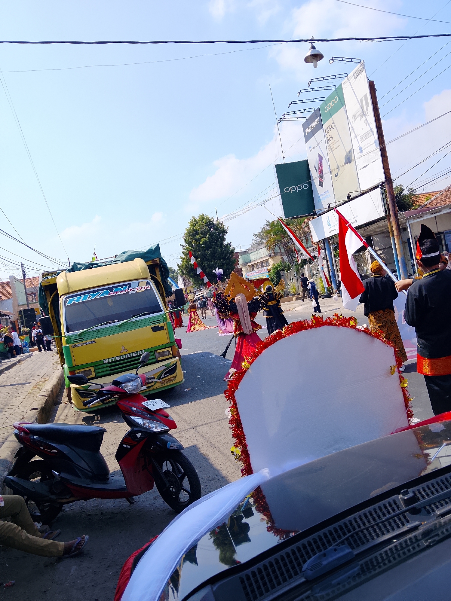 The image captures a street scene in Kertosono, Indonesia, possibly during a parade or festival. In the foreground, the hood of a dark-colored vehicle, likely a car, is visible, with its windshield wipers and defroster vents apparent. To the left, a red and black Yamaha scooter is parked on the side of the road, with a person's legs and feet visible as they sit on the curb.
Dominating the mid-ground is a bright yellow truck with green accents, featuring the text "RIZAL PRODUCTION" on its windshield. The truck appears to be part of a procession, with people in traditional attire and Indonesian flags visible on the street. Men in black tops and patterned waistcloths, some wearing hats, are seen holding flags. Women in elaborate, colorful costumes, adorned with golden headdresses, are also present.
In the background, there are buildings, trees, and overhead power lines. A prominent billboard advertising "SAMSUNG" and "OPPO" phones is visible on the right side of the street, alongside a street lamp. The sky is clear and blue with some scattered clouds. The overall impression is one of a lively, outdoor event taking place in an urban environment.