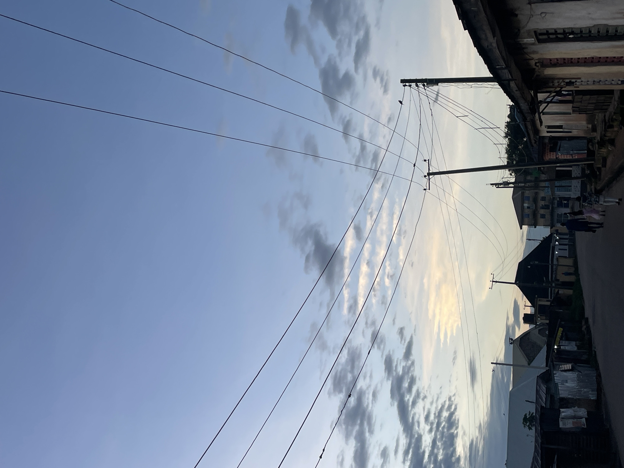 The image depicts a street scene in Jos, Nigeria, at dusk. The sky is a pale blue with scattered clouds, illuminated by the setting sun. Several power lines stretch across the frame, supported by wooden poles. On the right side of the street, there are a row of buildings with corrugated metal roofs and some with concrete facades. Some of these structures appear to be small shops or residences. On the left side of the street, more buildings are visible, including some with pitched roofs and what appears to be a water tank on one of them. Further down the street, there are multi-story buildings. Two people, one adult and a child, are walking away from the camera down the street. A single bird is perched on a power line. The overall impression is of a typical street in a Nigerian city during the transition from day to night.