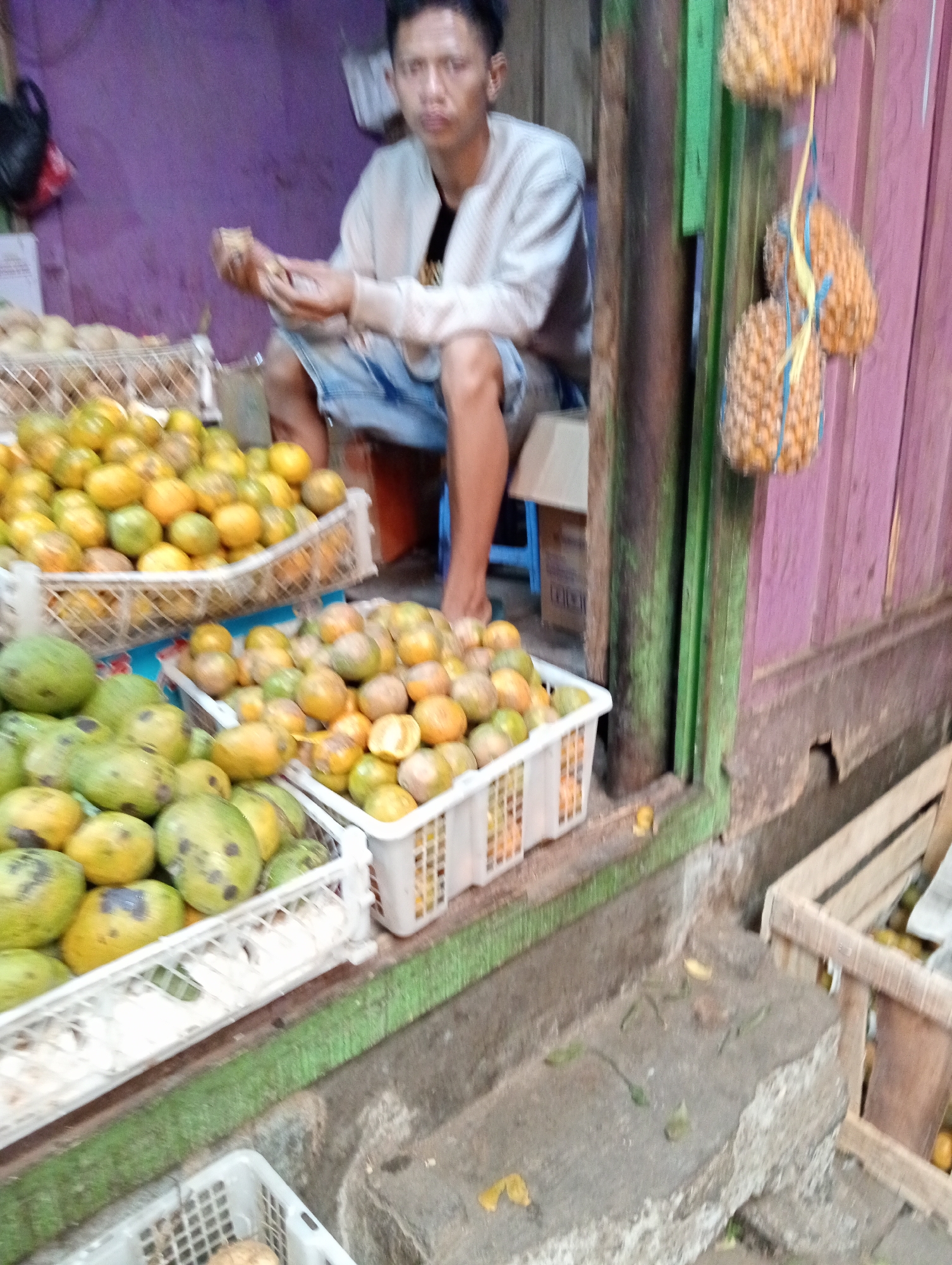 This is a medium-shot, eye-level, horizontal photograph of a street fruit stand in Grabag, Indonesia. In the background, a young man is seated behind a display of fruits. He is wearing a light-colored long-sleeved shirt and denim shorts. He is holding a piece of fruit in his hands, seemingly inspecting it. The fruit stand features several baskets filled with oranges and mangoes. Hanging from a wooden beam to the right are three small pineapples, tied with blue string. The background shows a purple wall and a wooden structure with green trim. The foreground depicts a weathered concrete step and a wooden crate filled with more fruit. The overall lighting is dim, suggesting it might be late afternoon or a shaded area.