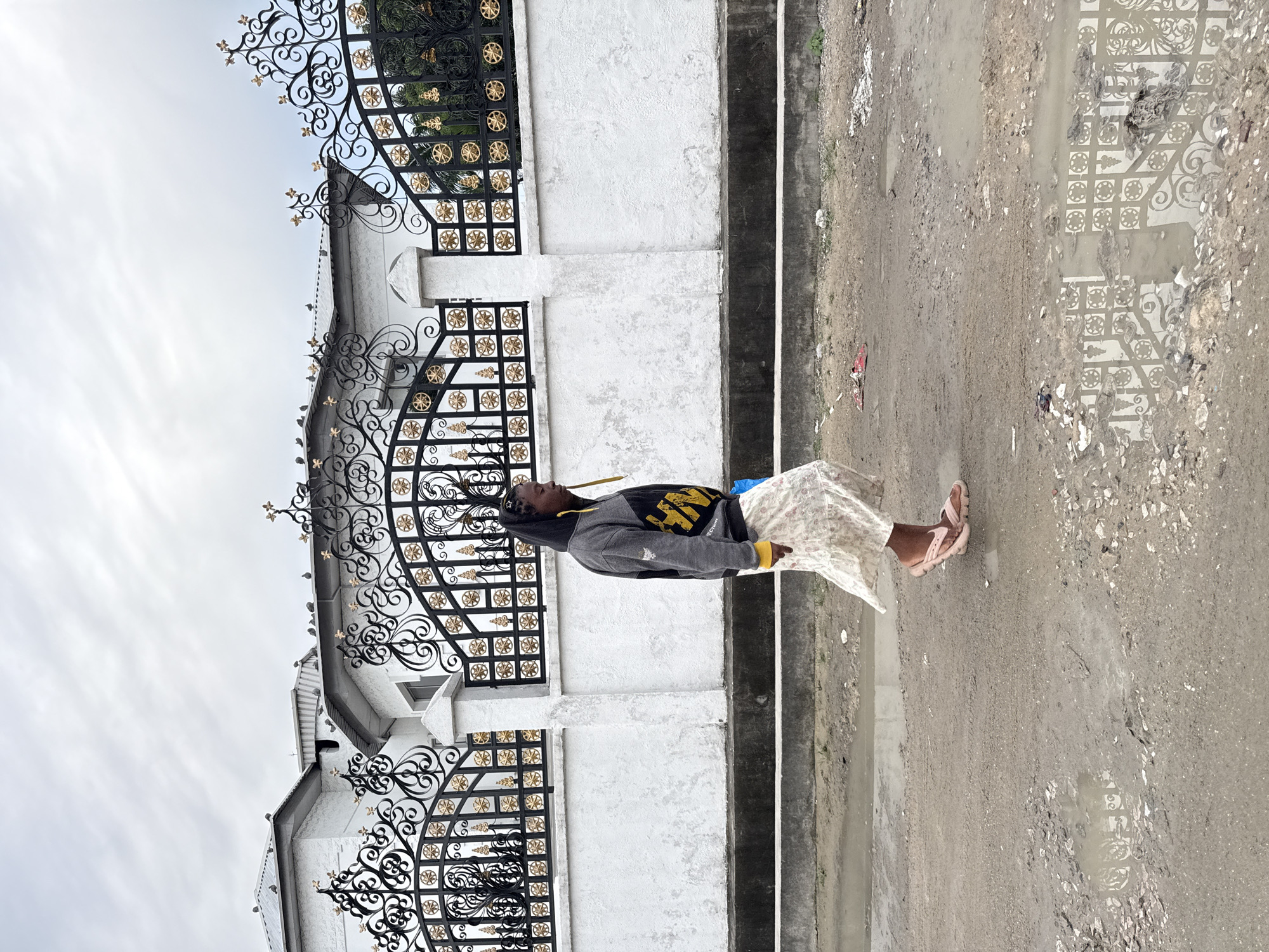 A person stands in front of a white, gated structure. The person is wearing a grey hoodie with yellow accents, a light-colored skirt, and pink sandals. The gate is black with gold decorative elements. The ground is a mix of dirt and pavement with puddles of water. The reflection of the gate can be seen in the puddles. The sky is overcast. The scene appears to be in Ado, Nigeria.
