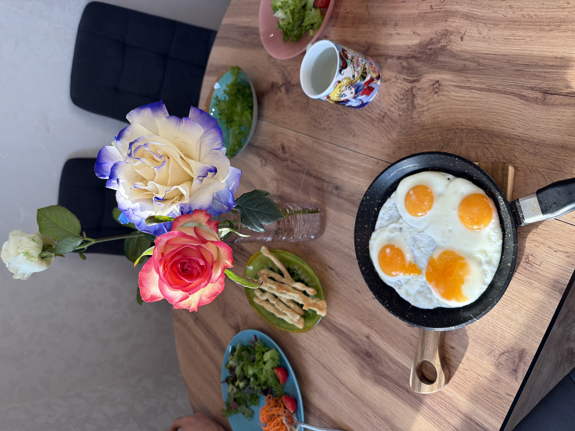 A high-angle shot shows a wooden table in Izhevsk, Russia, set with a partially visible black chair to the left. A vase of roses, including one with white petals edged in blue, sits beside a small plate of salad, a patterned mug, another small plate of salad, a plate of breadsticks, and a frying pan containing four fried eggs. A person is visible in the lower left.